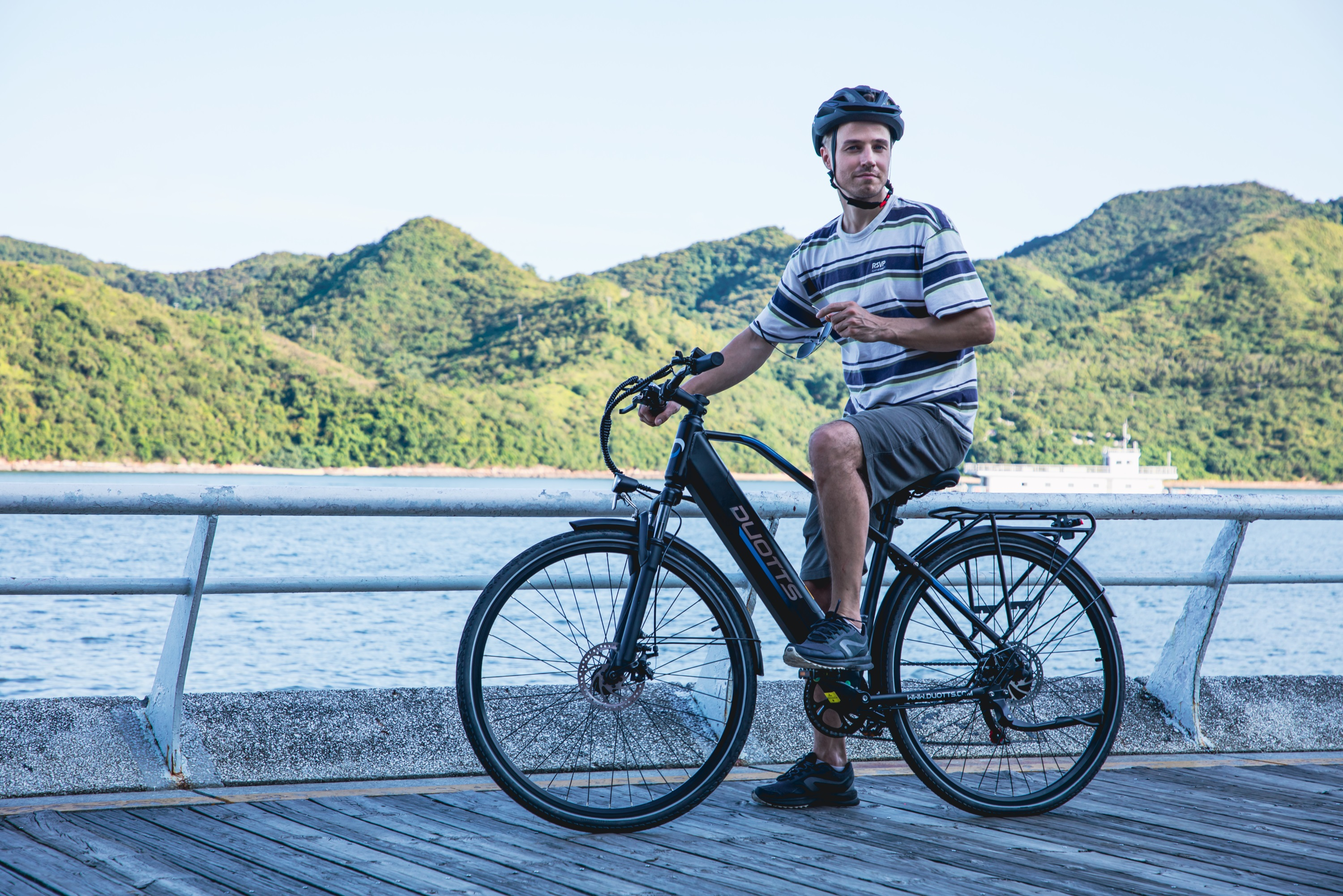 Hombre con casco en e-bike en muelle con colinas verdes y agua.