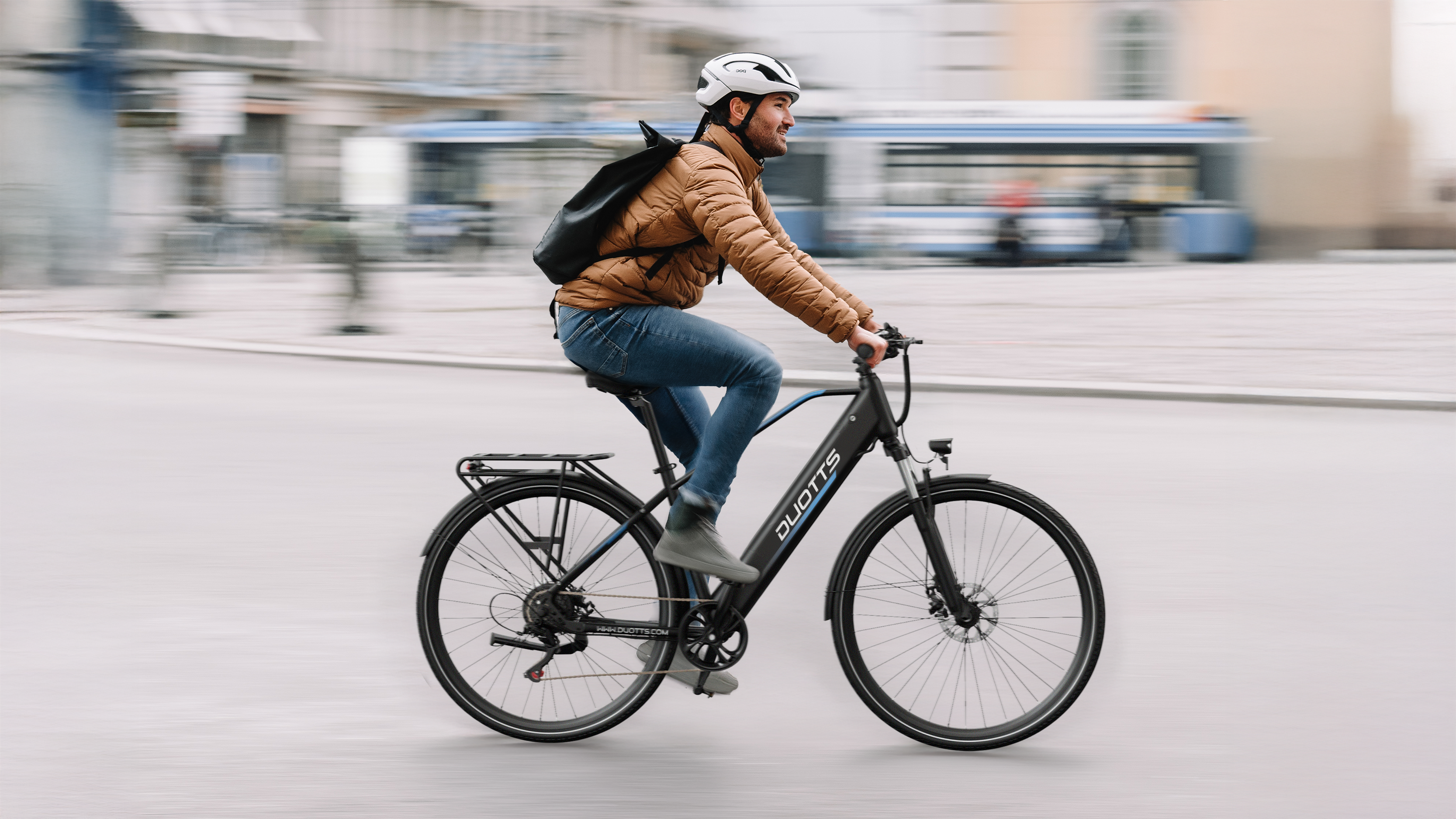 Hombre en bicicleta eléctrica con chaqueta marrón, jeans azules, casco blanco.
