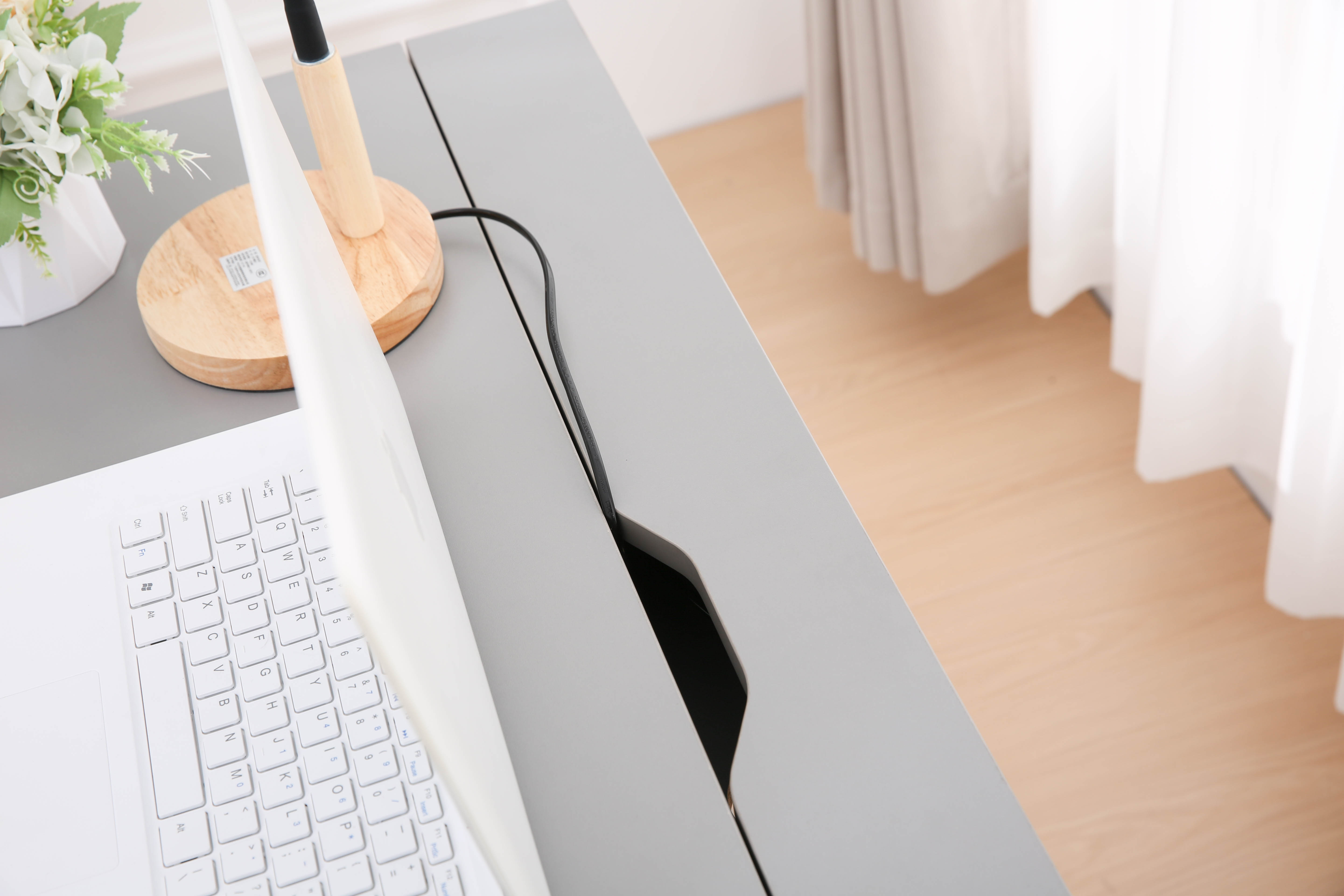 Biały laptop na szarym biurku z lampą i kwiatami. Widoczny jest kabel zasilający.