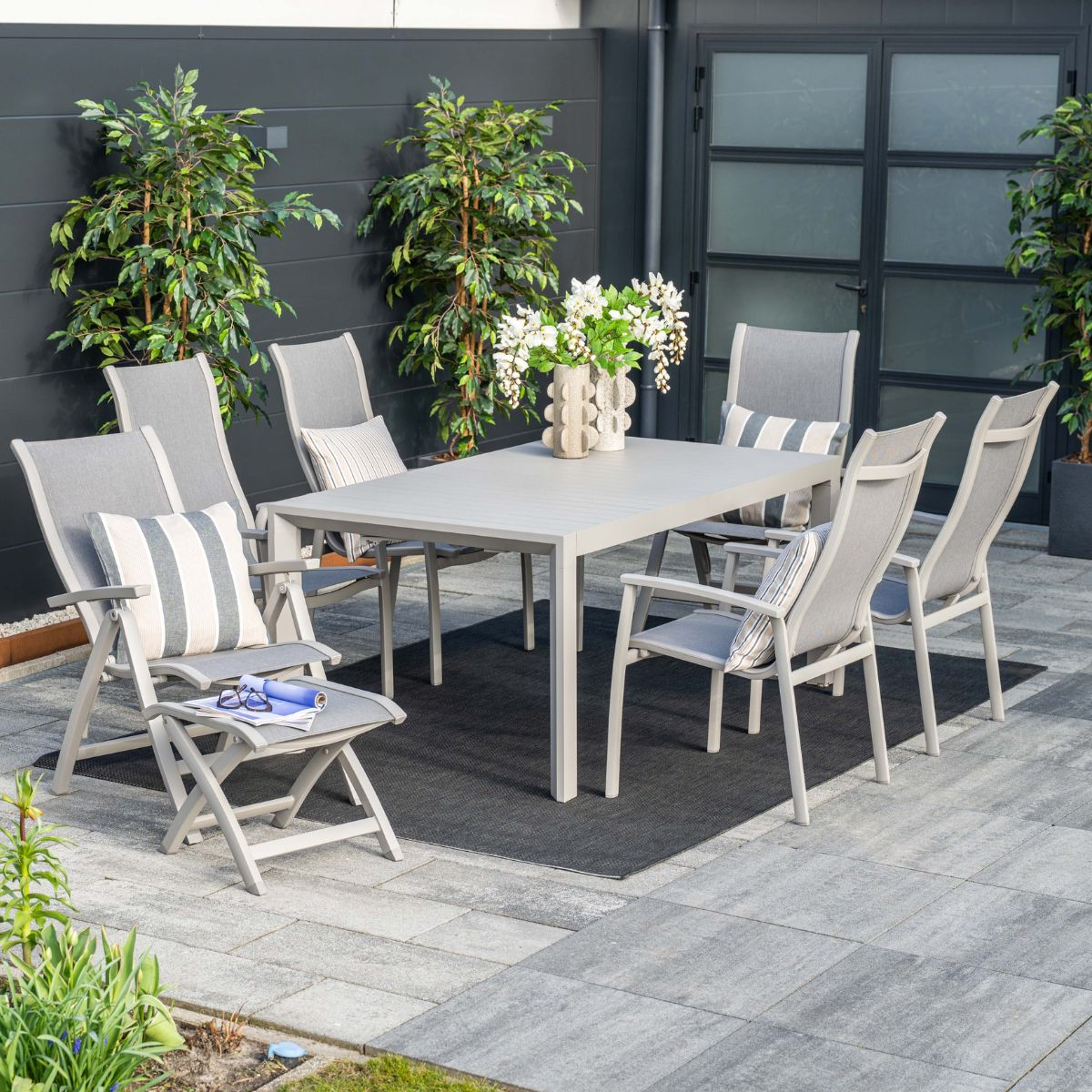 Grijze buitentafel met zes stoelen op een betegeld terras, versierd met kussens en planten.