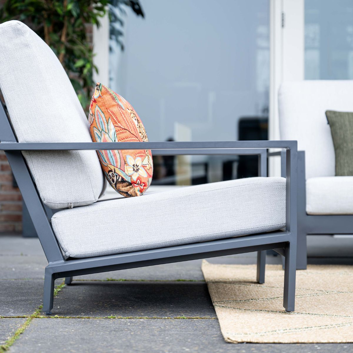 Grijze fauteuil met kleurrijk kussen op stenen patio met vloerkleed.