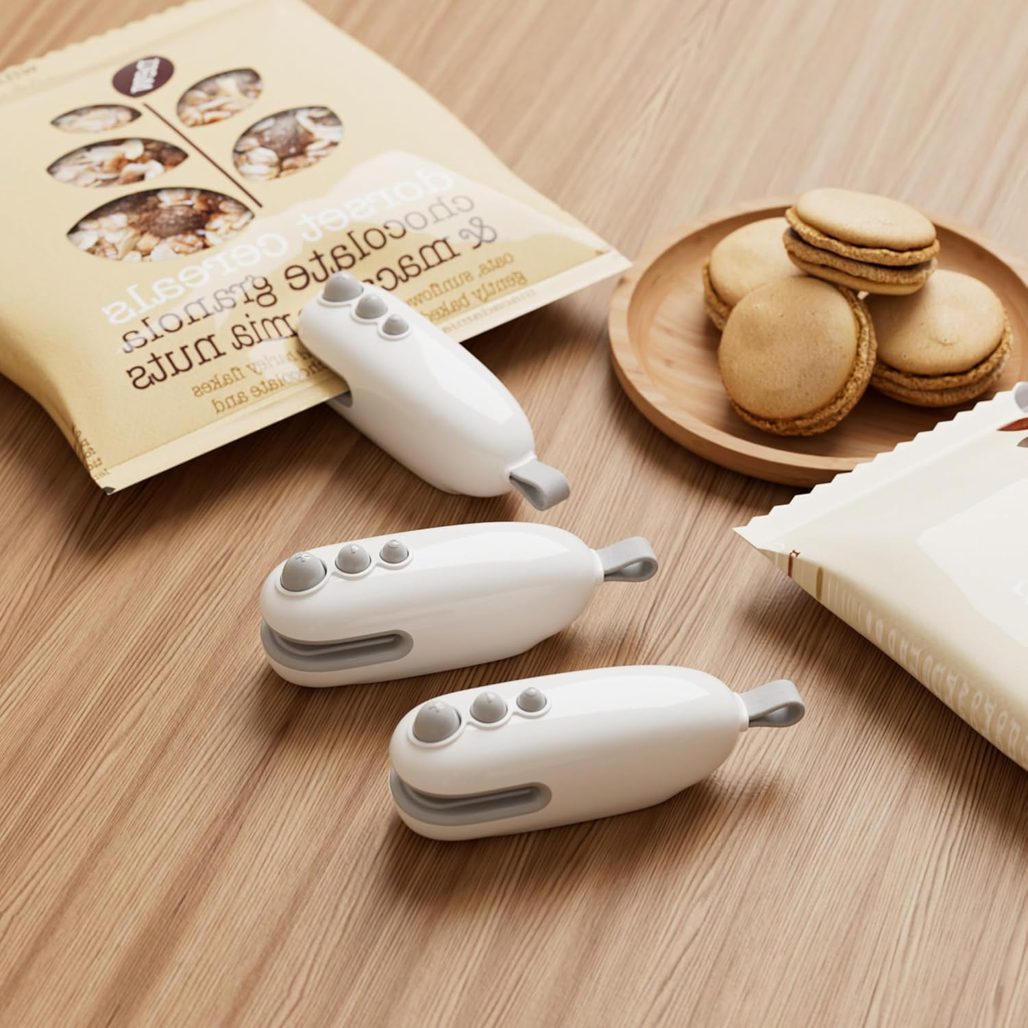 Drei weiße Beutelclips mit grauen Akzenten liegen auf einem Holztisch neben Keksen.