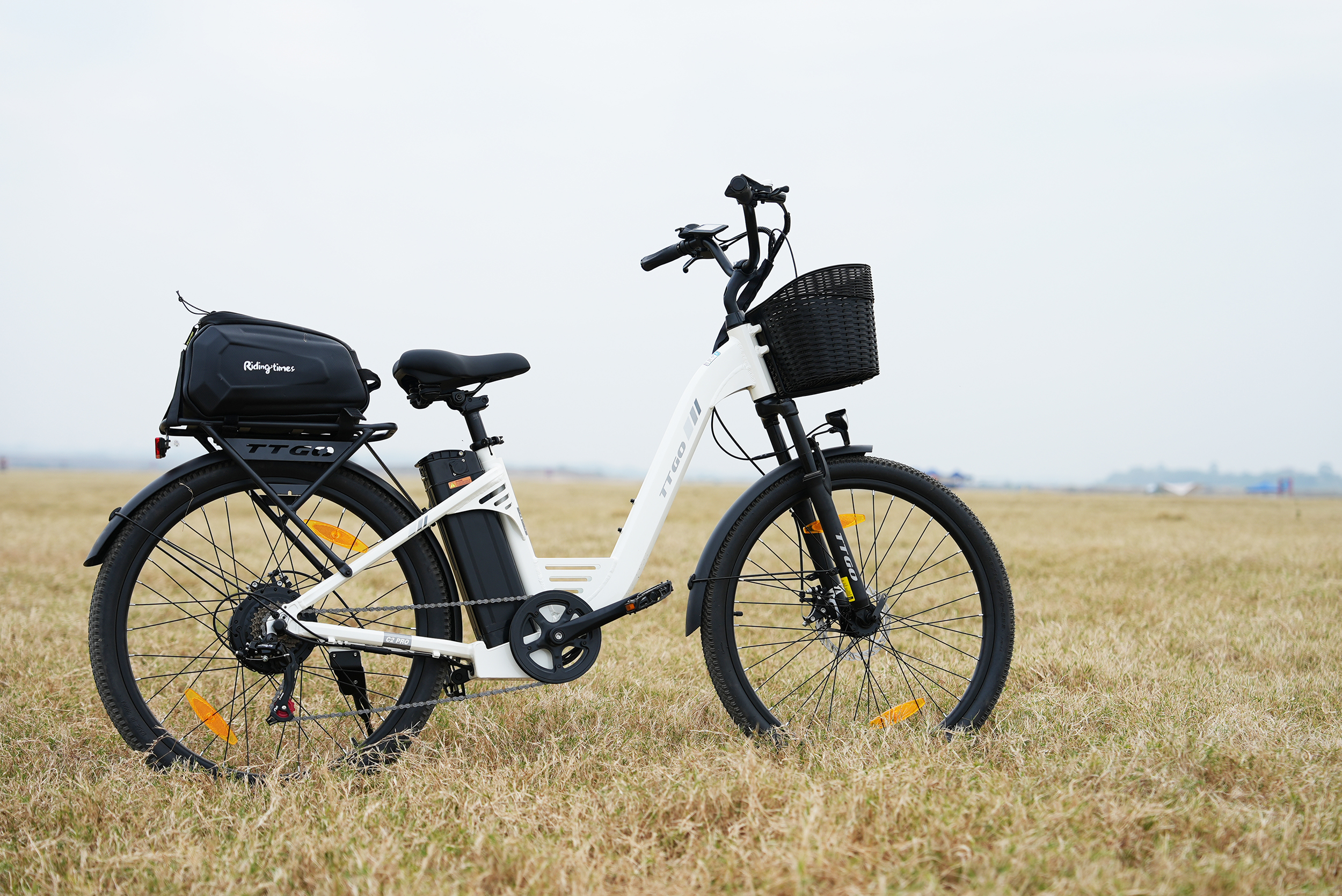 Ein weißes Elektrofahrrad mit schwarzem Korb und Gepäckträgertasche auf einem Feld.