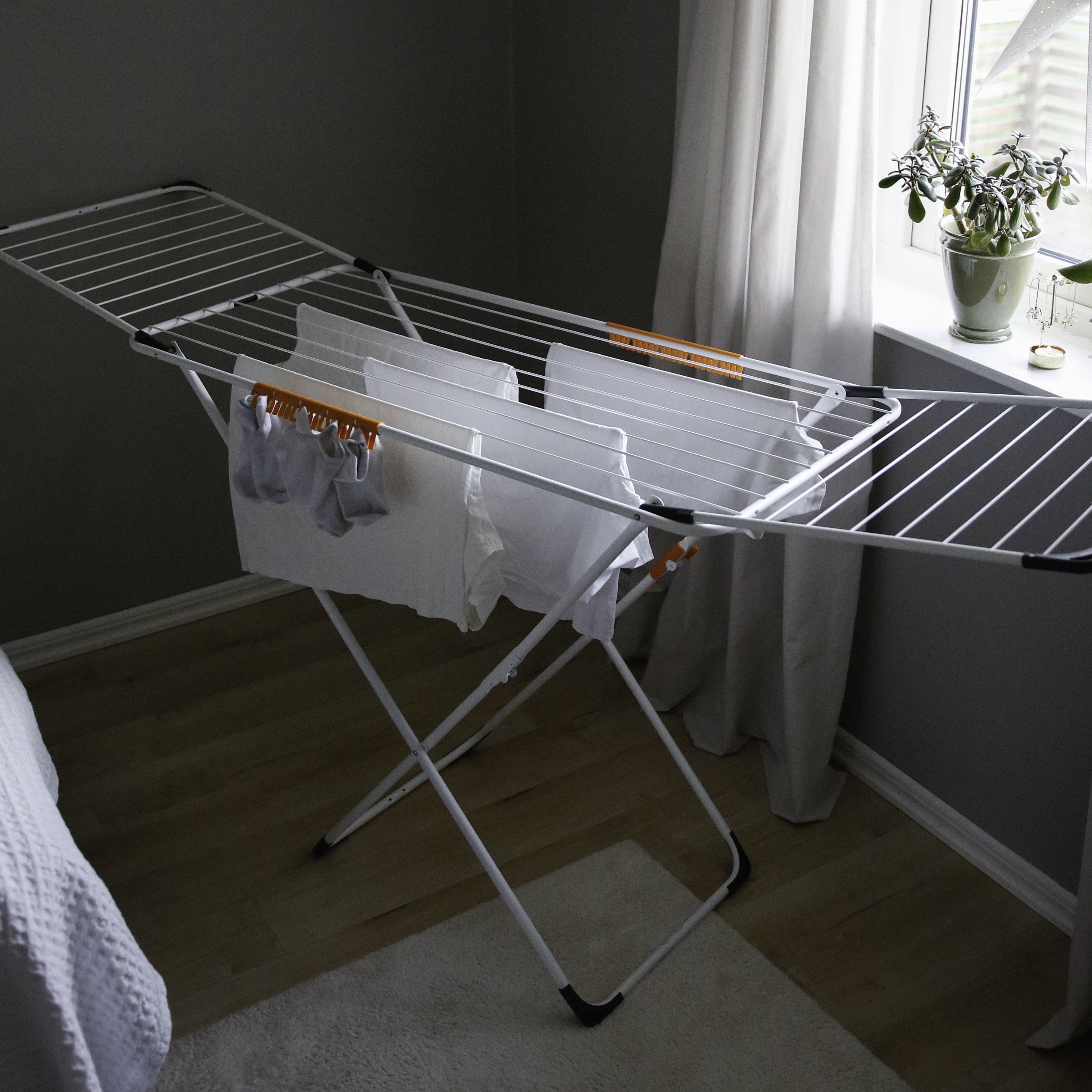 A white drying rack with clothes stands on a wooden floor next to a window.