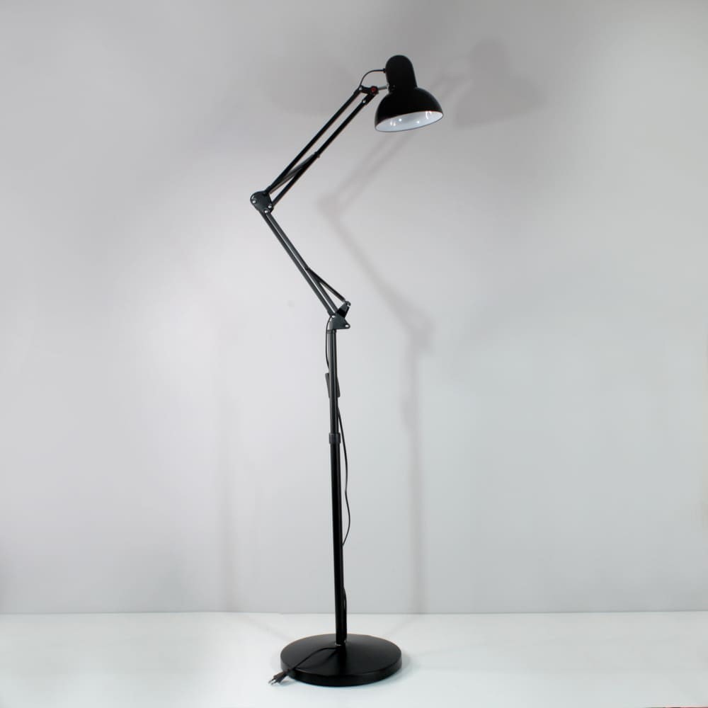 Una lámpara de pie articulada negra está sobre una superficie blanca contra una pared gris.