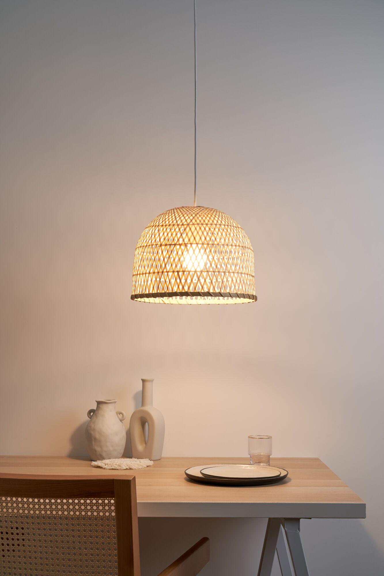 Okrągły klosz lampy wiszącej z wikliny oświetla drewniany stół z dwoma wazonami i naczyniami.