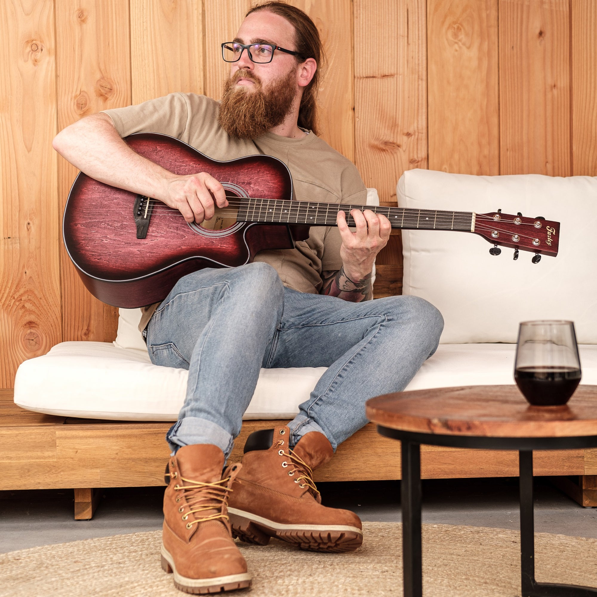 Man met baard en bril speelt gitaar op bank, houten lambrisering achtergrond.