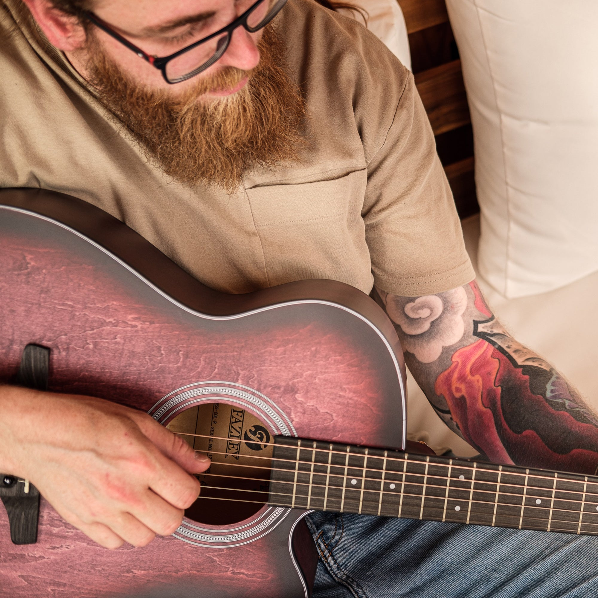 Man met baard speelt gitaar. Hij draagt een t-shirt en spijkerbroek. Zijn arm is getatoeëerd.