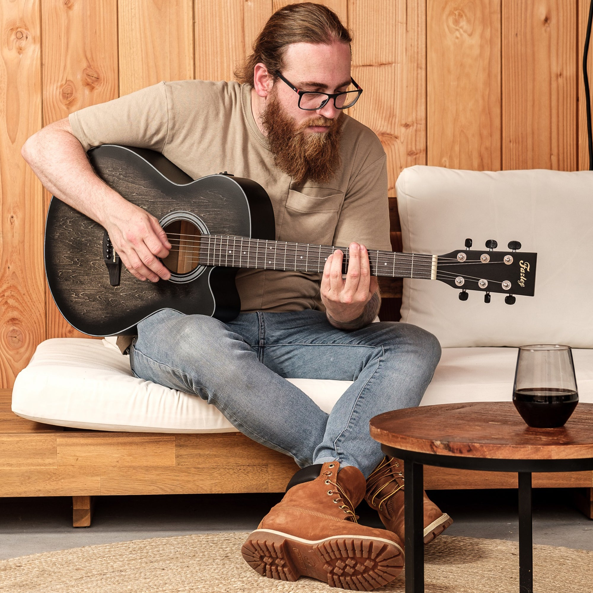 Man met baard speelt akoestische gitaar op bank, houten lamellen achtergrond.
