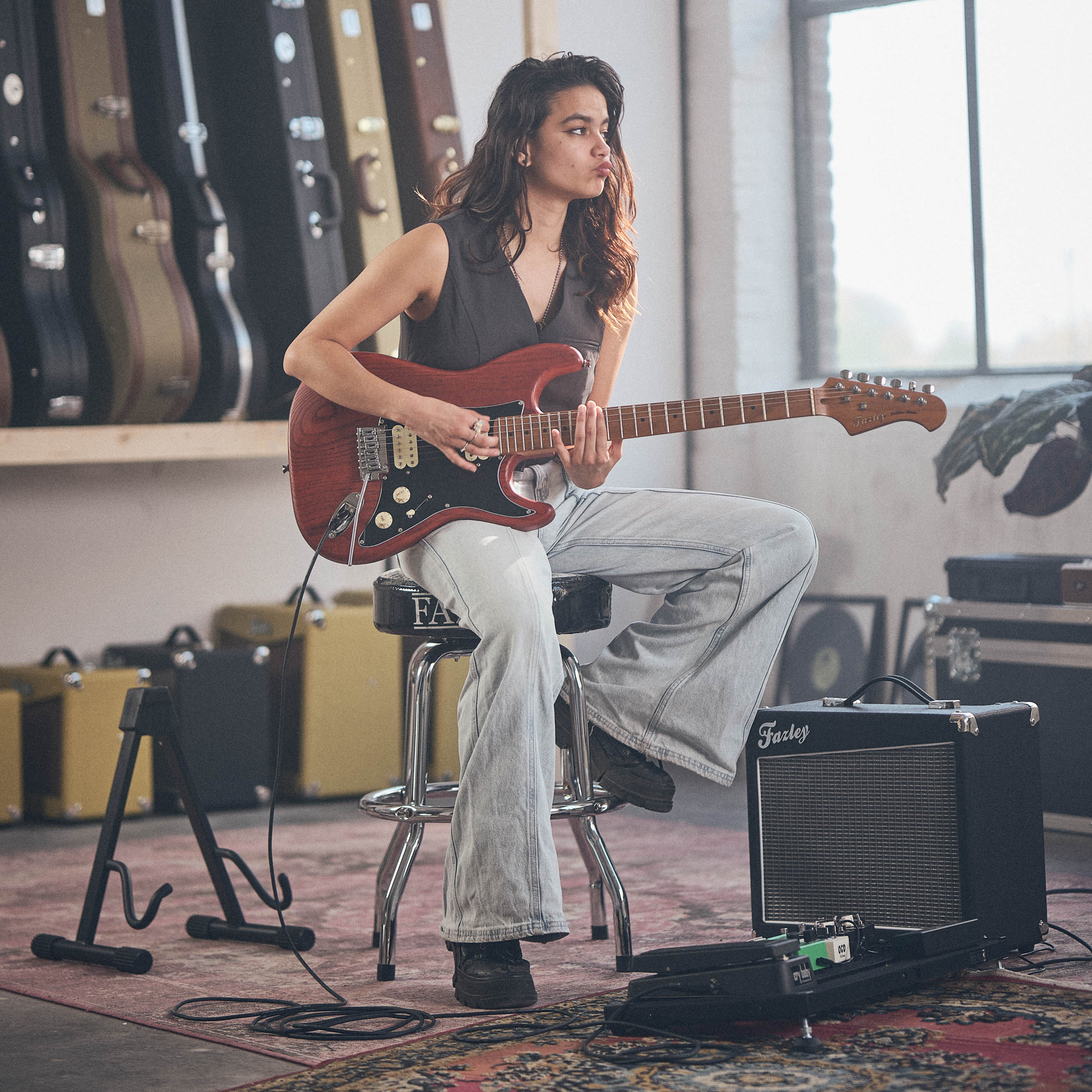 Een vrouw zit op een kruk en speelt gitaar in een ruimte vol gitaarkoffers.