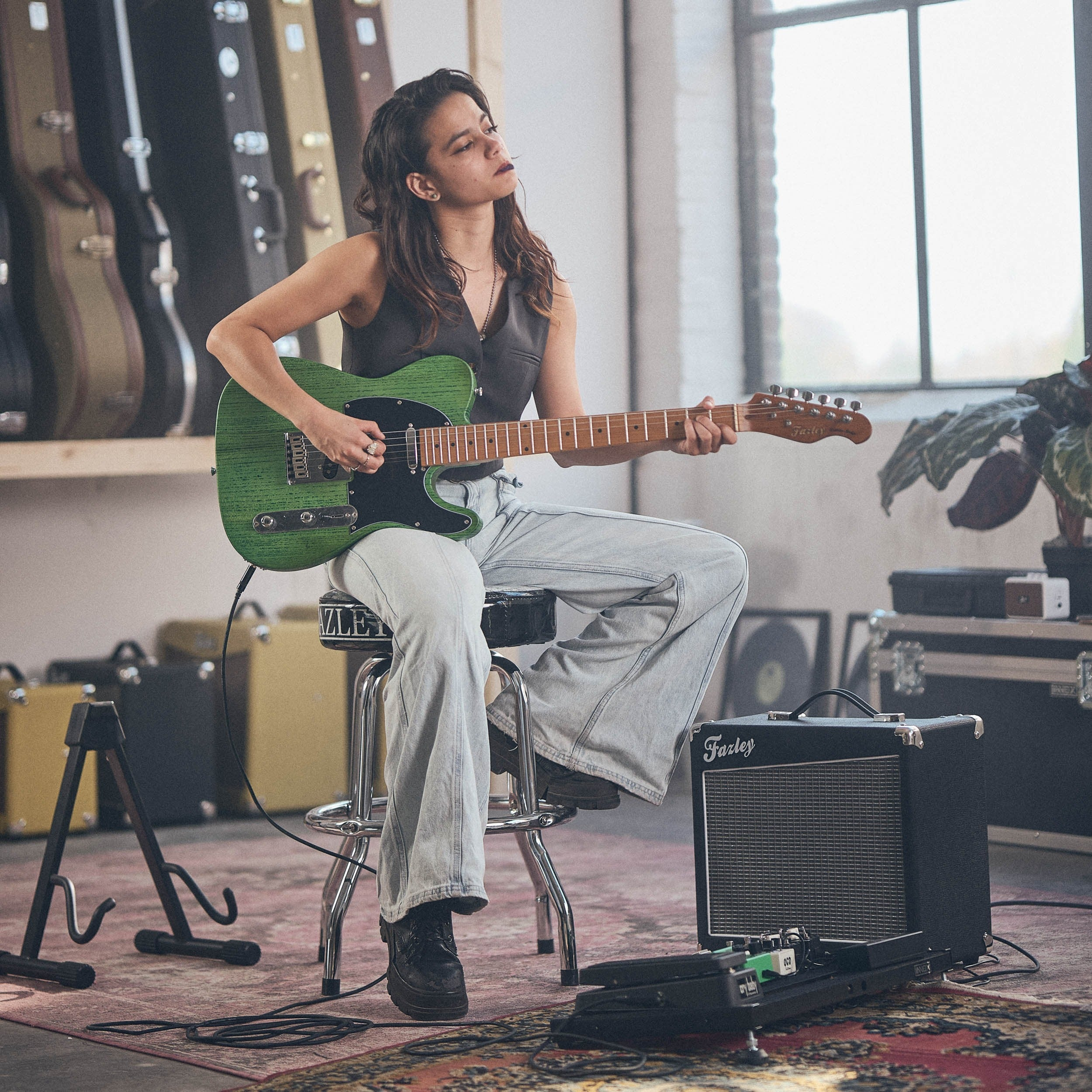 Vrouw speelt groene elektrische gitaar, zit op kruk, versterker en gitaarkoffers op achtergrond.