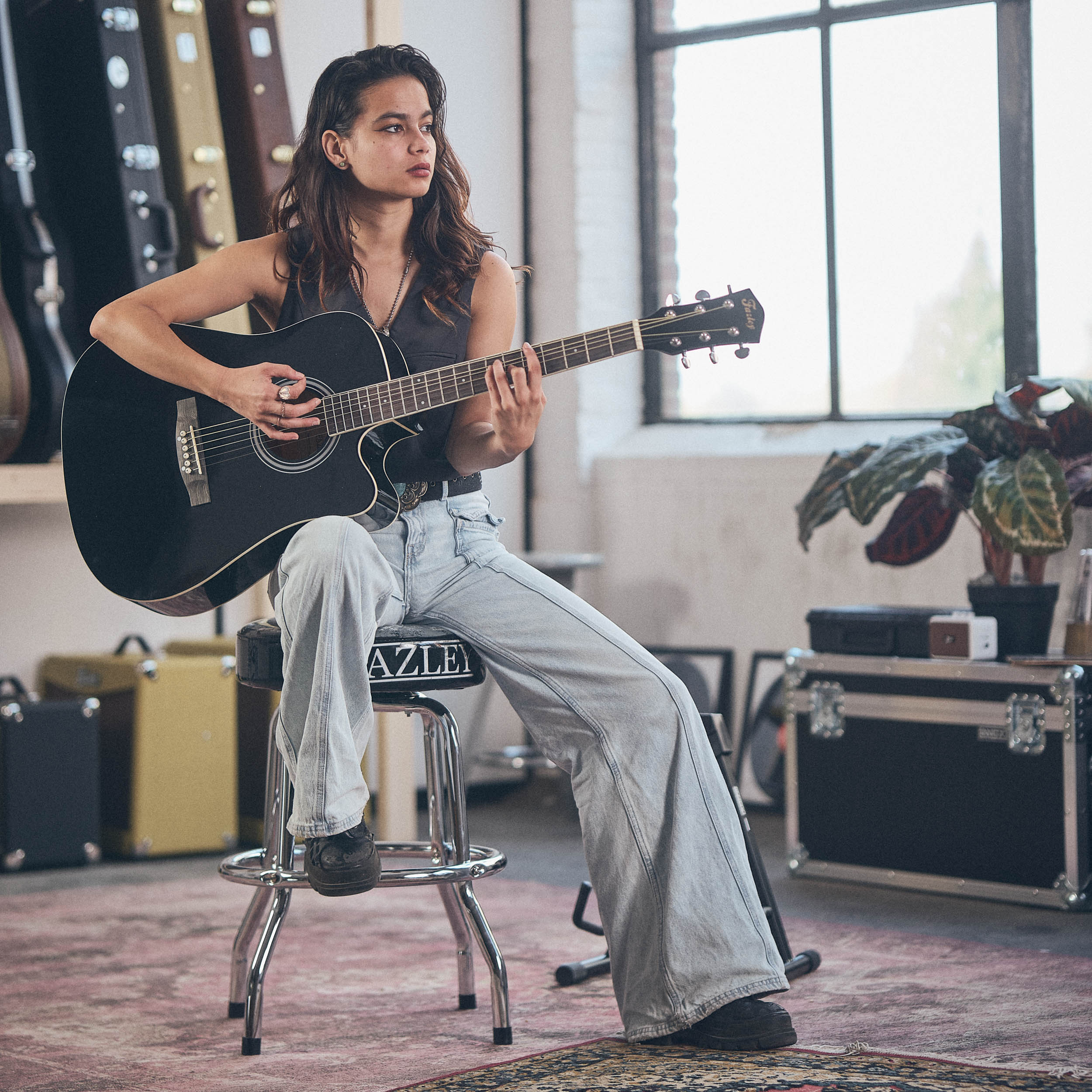 Een jonge vrouw met lang haar speelt op een zwarte akoestische gitaar, zittend op een kruk.
