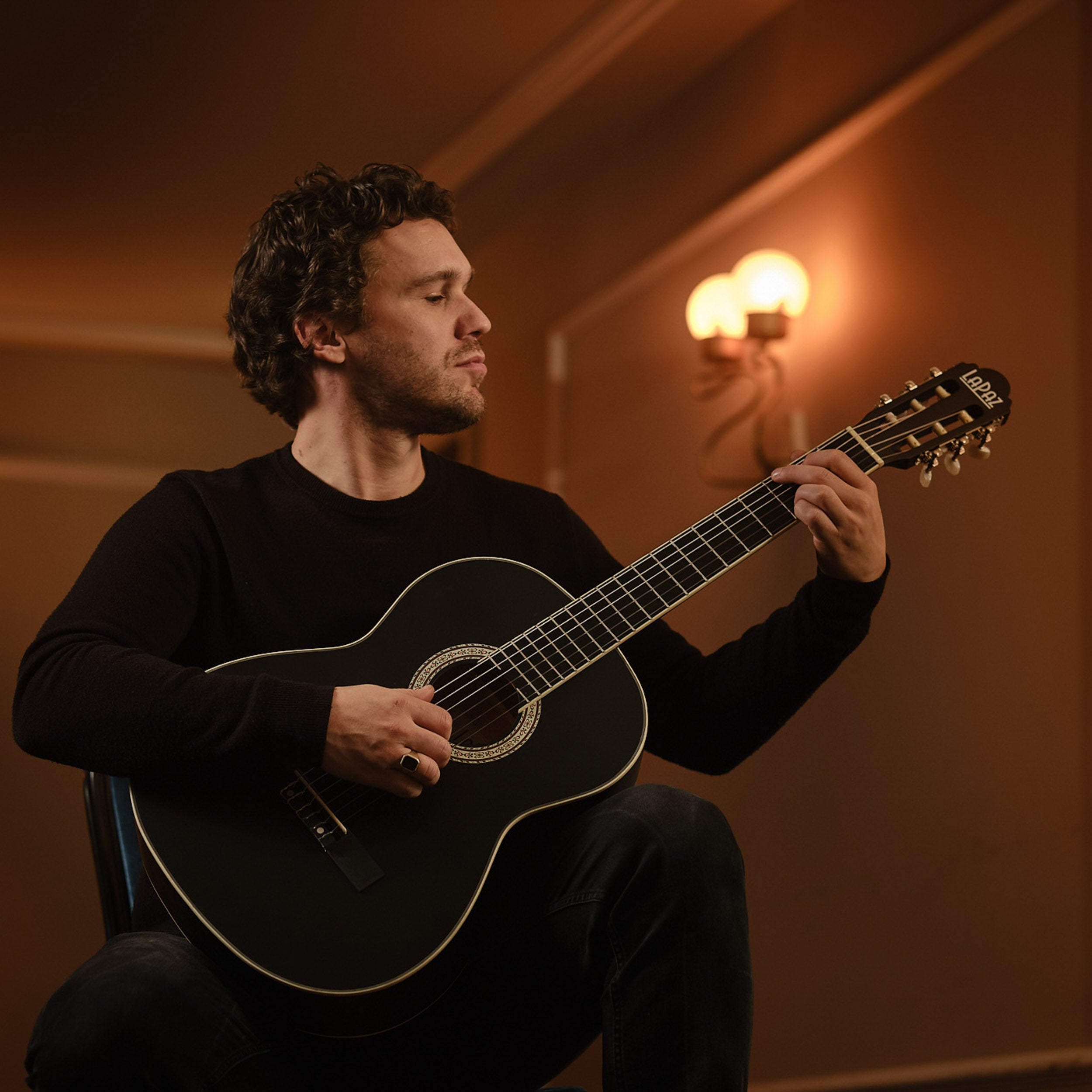 Een man in een zwart trui speelt op een zwarte gitaar. Warme, sfeervolle verlichting.