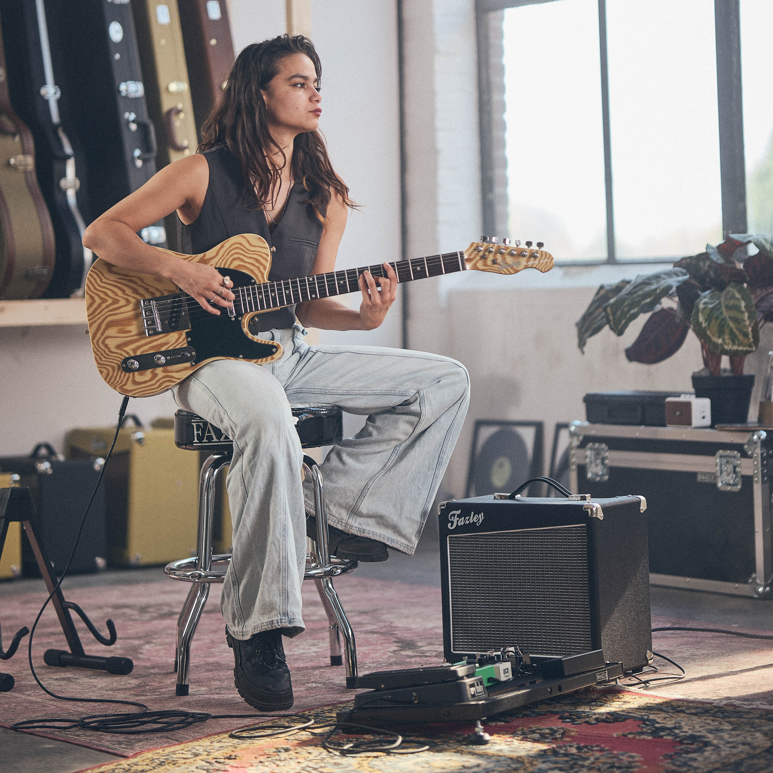 Een vrouw zit op een kruk gitaar te spelen in een kamer met gitaarkoffers.