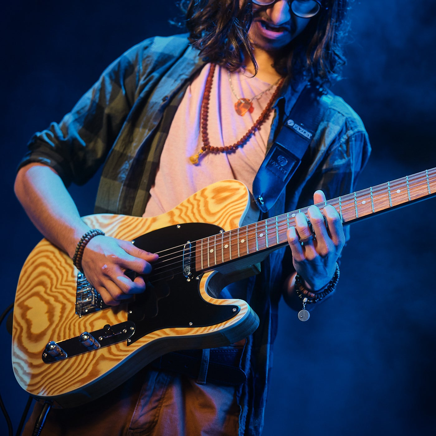 Een man bespeelt een lichtgekleurde elektrische gitaar in blauw licht. Hij draagt kralenkettingen.