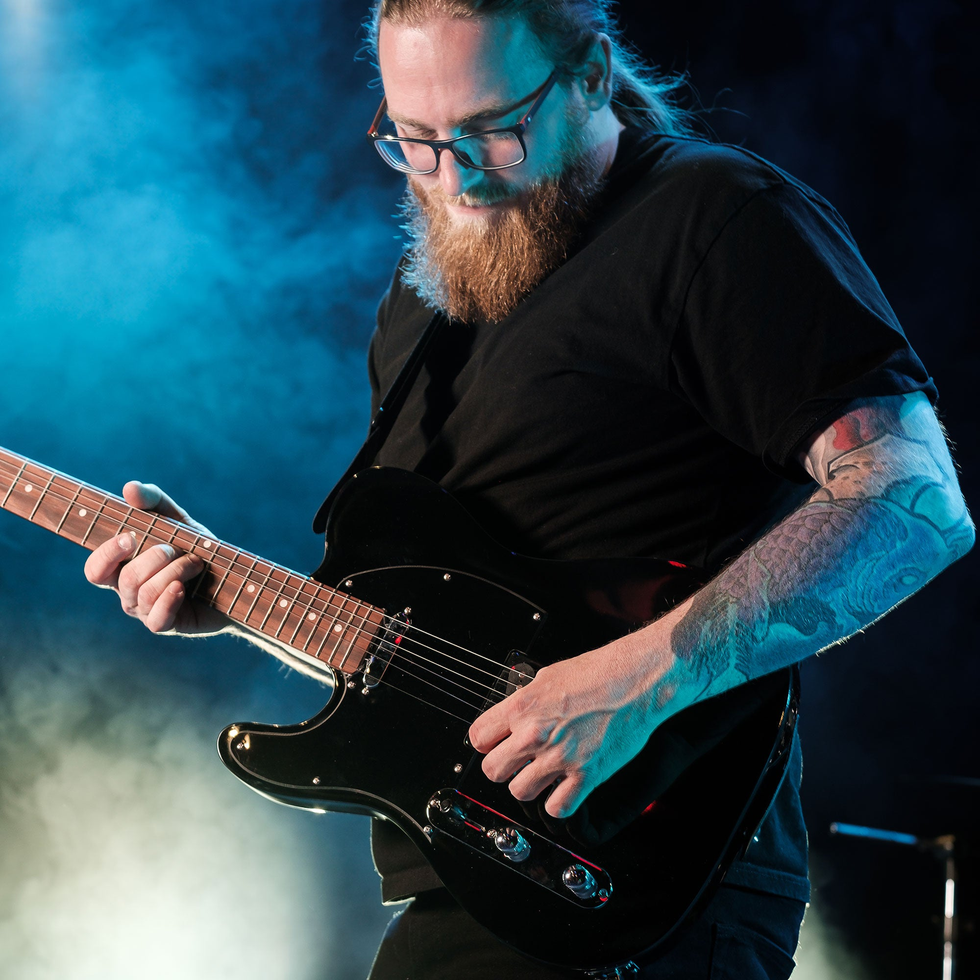 Man met bril en baard speelt elektrische gitaar op podium met blauwe rook.
