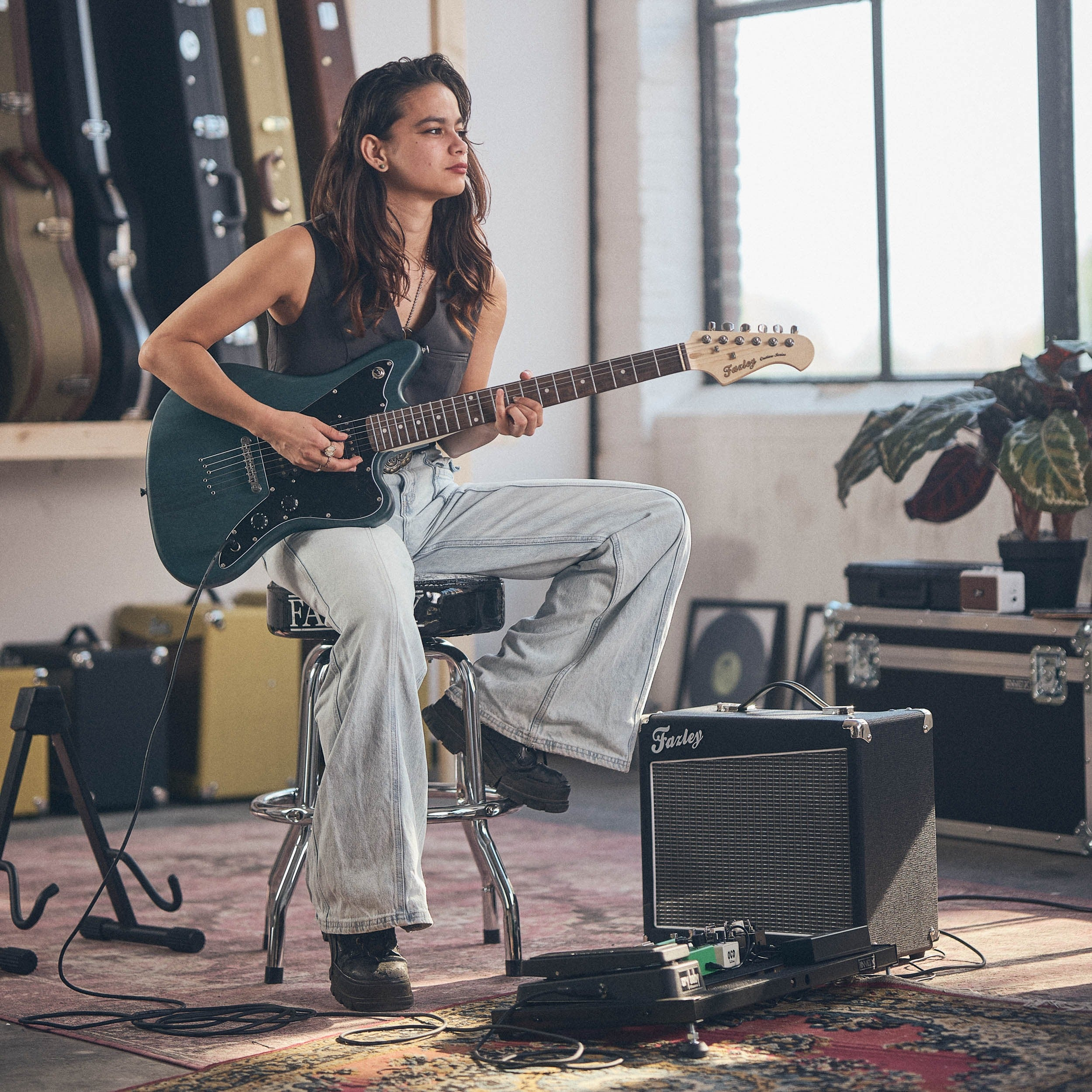 Een vrouw zit op een kruk en speelt op een groene elektrische gitaar in een kamer met gitaarkoffers.