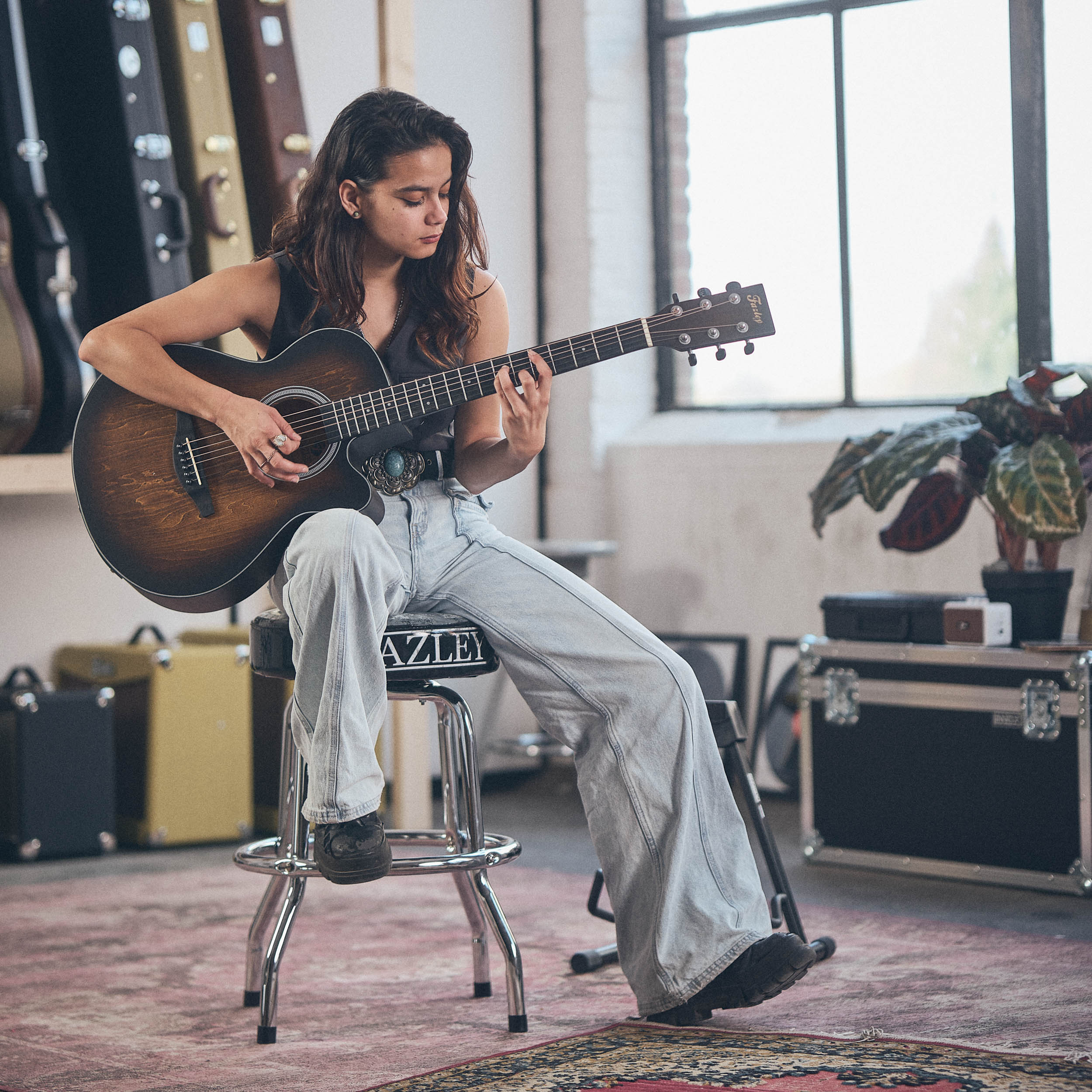 Een vrouw zit op een kruk een gitaar te spelen in een kamer met gitaarkoffers.