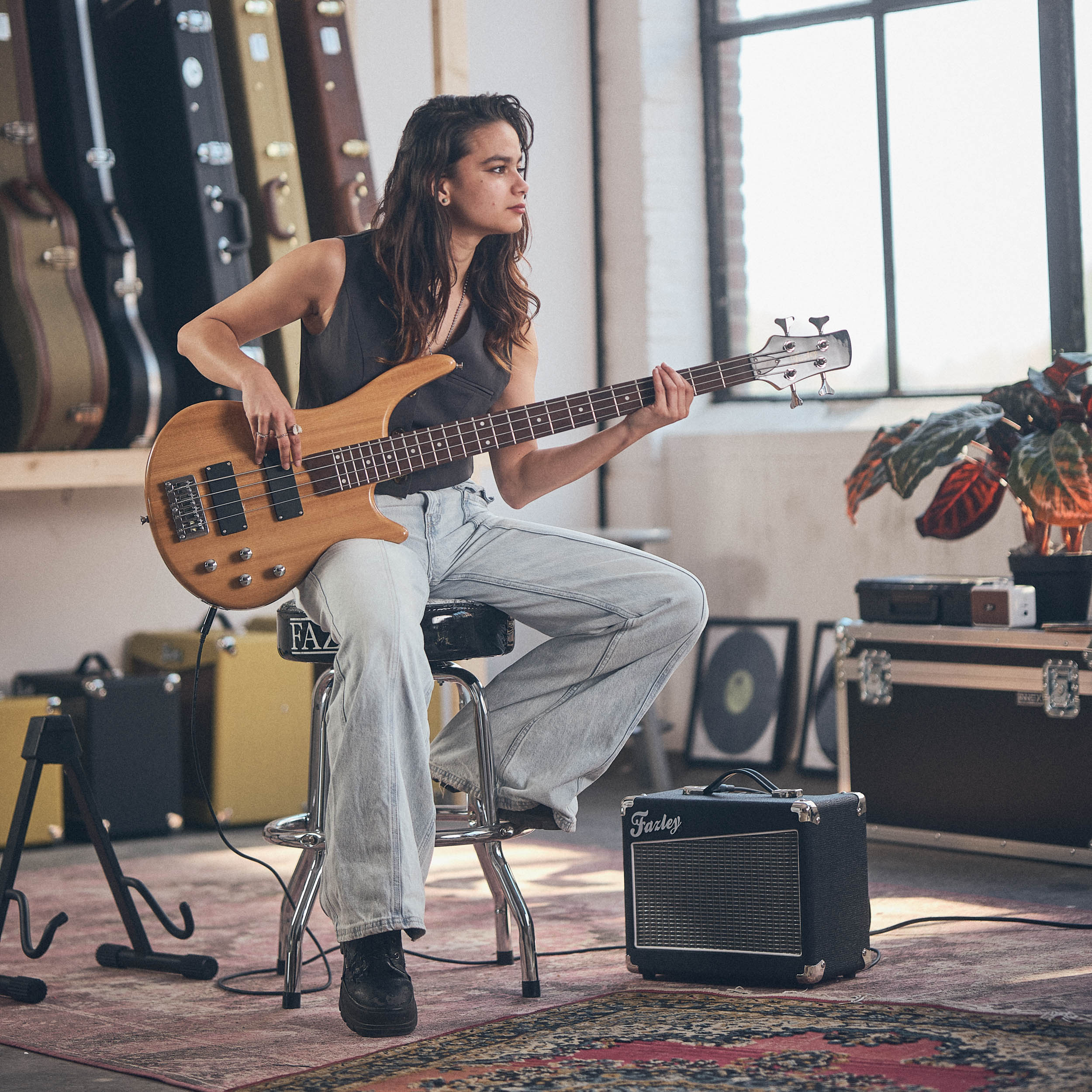 Een vrouw met lang haar speelt basgitaar, zittend op een kruk, gitaarkoffers op de achtergrond.