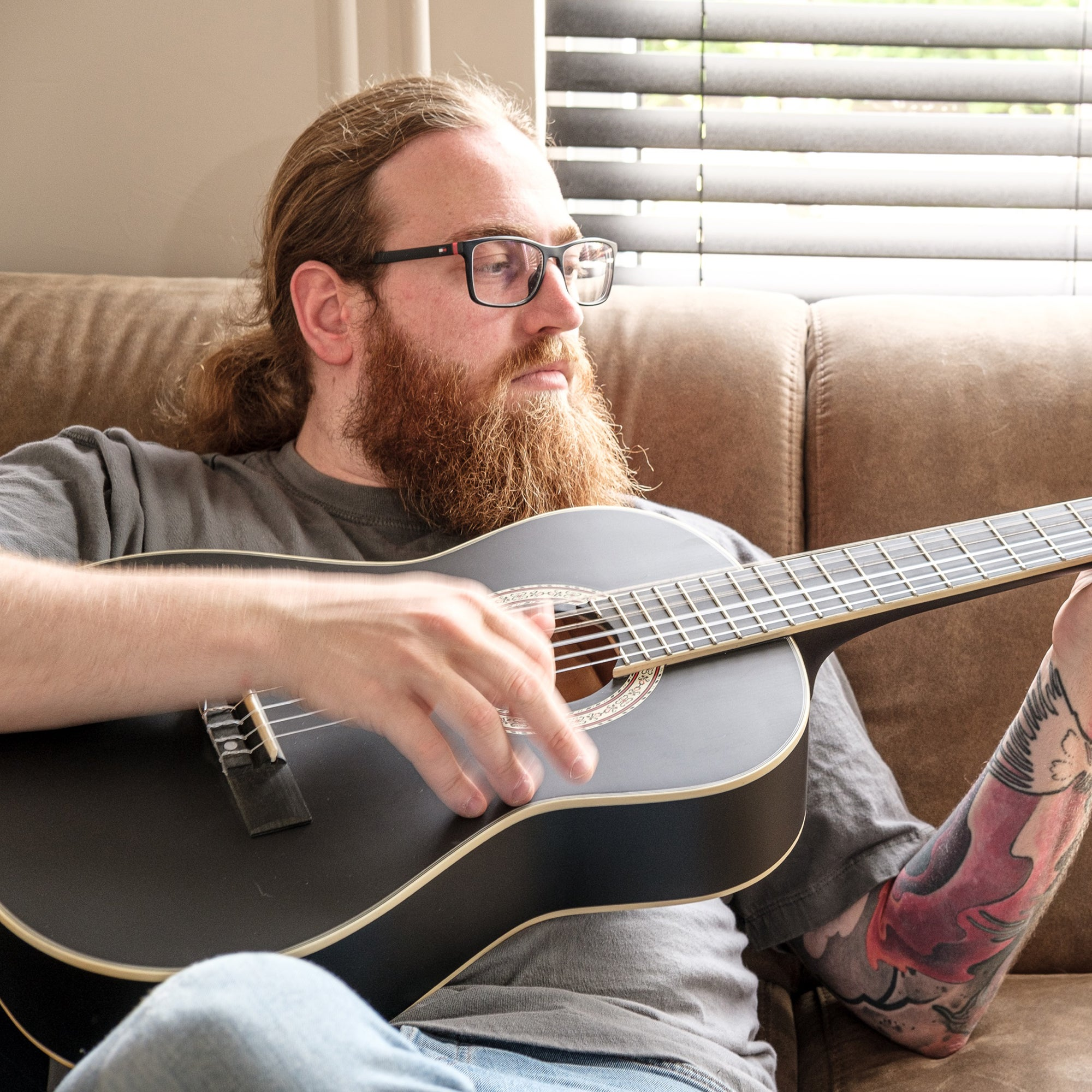 Een man met baard en bril speelt gitaar op een bruine bank.