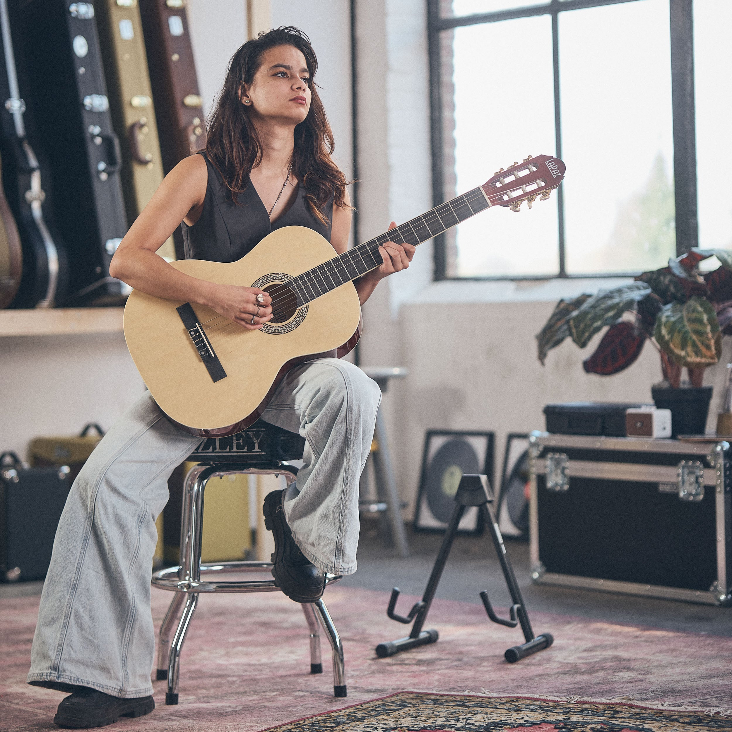 Een vrouw zit op een kruk en speelt gitaar. Gitaarkoffers staan op de achtergrond.