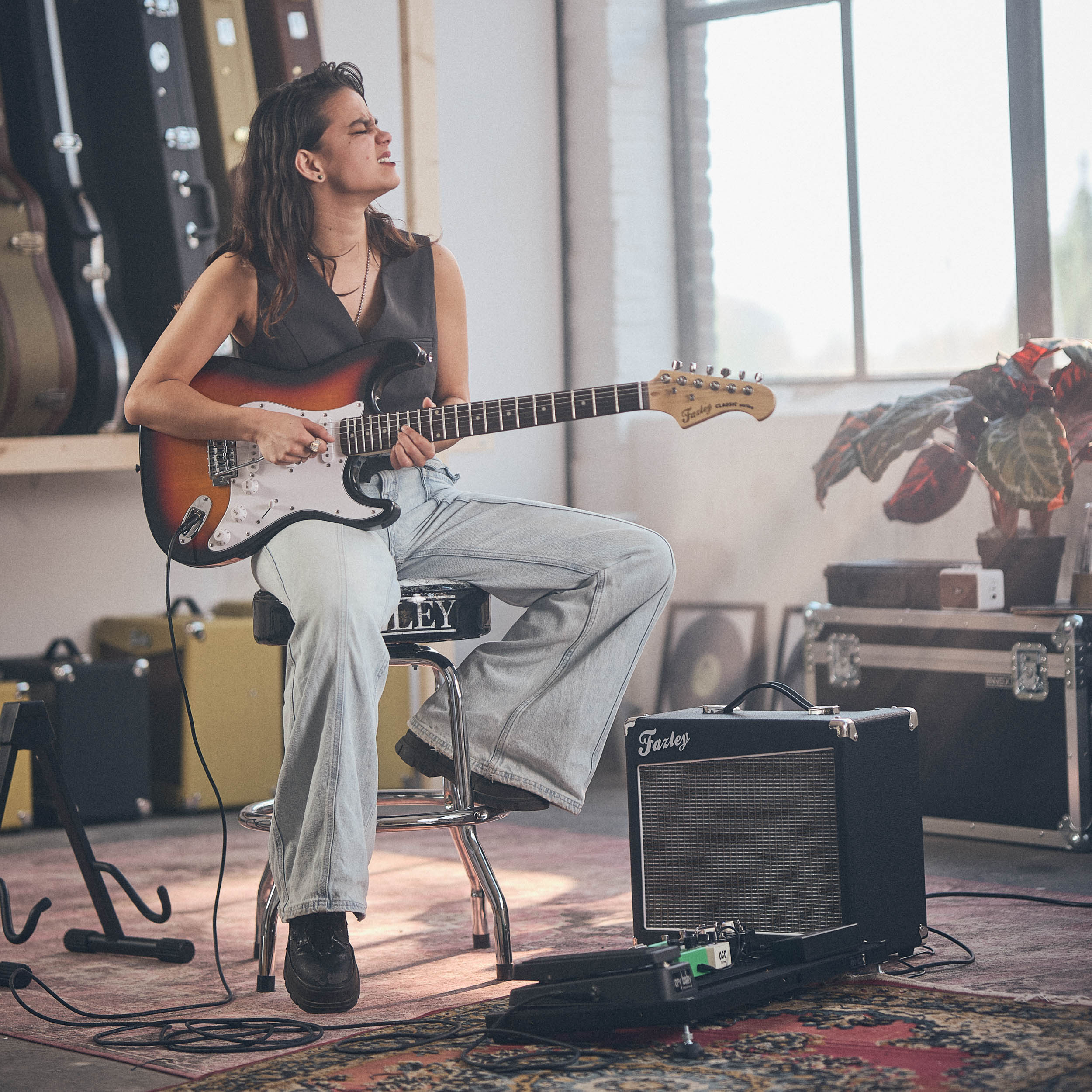 Vrouw speelt elektrische gitaar op kruk, versterker en gitaarkoffers op achtergrond.