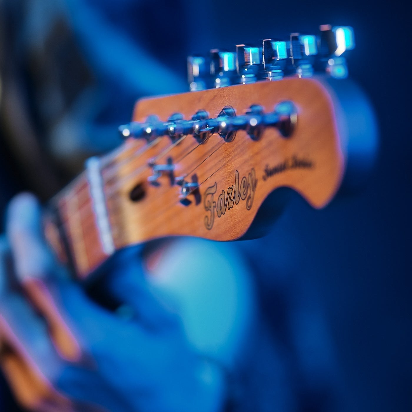 Close-up of a guitar headstock with tuning pegs and strings, illuminated by blue lights.