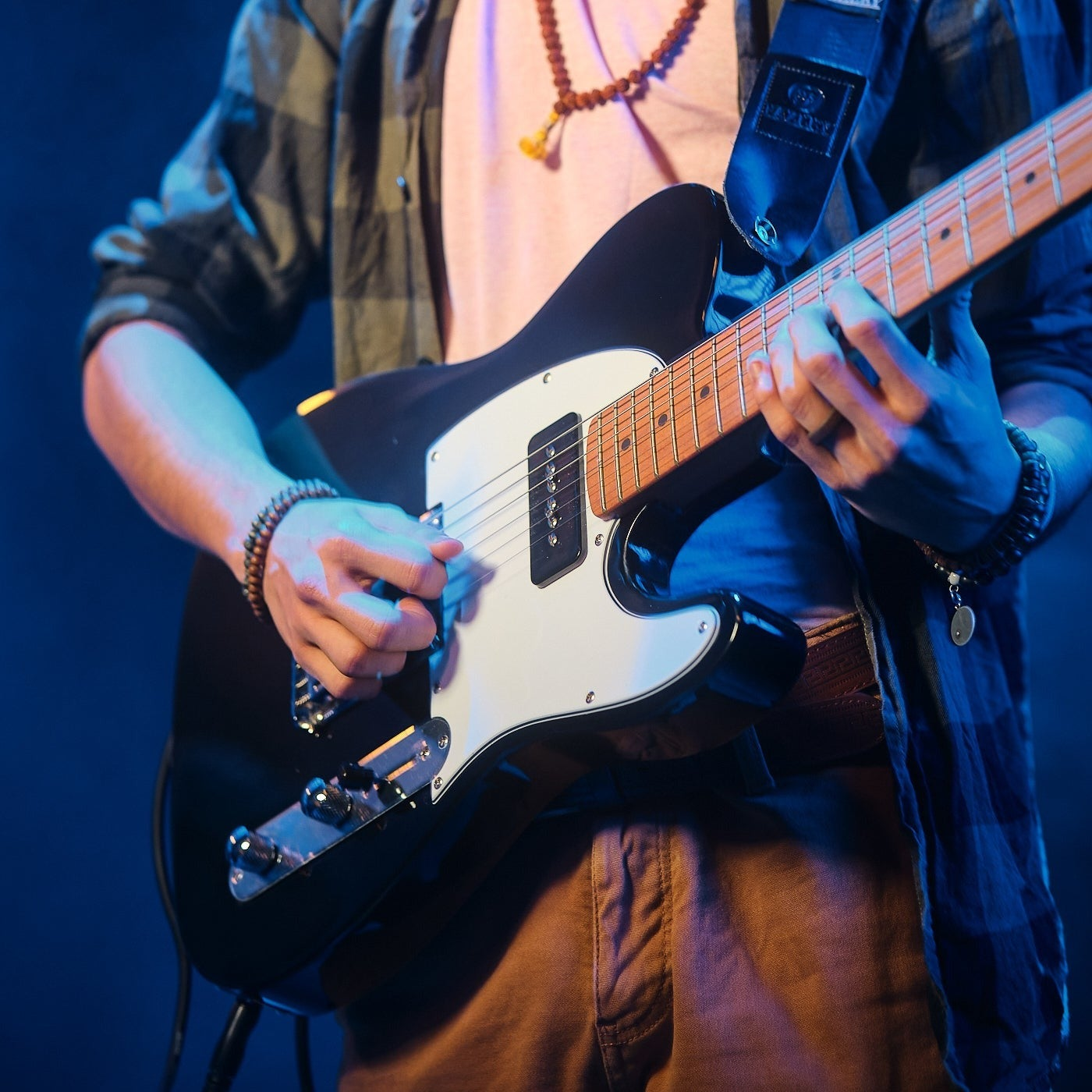 A person plays a black electric guitar, wearing brown pants and a checkered shirt.