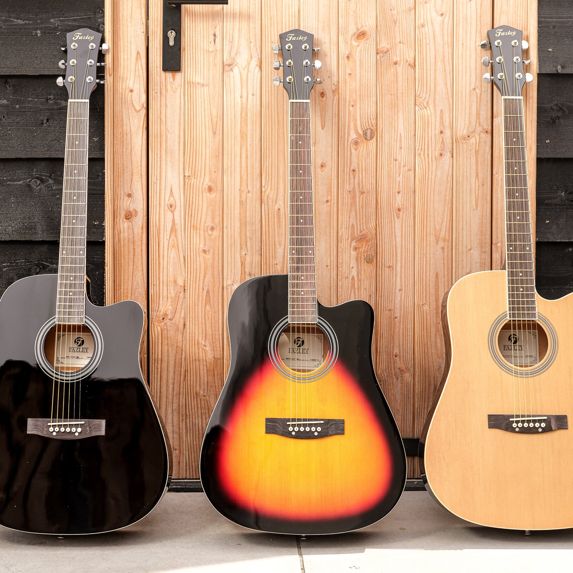 Drie akoestische gitaren voor een houten wand. Zwart, sunburst en naturel.