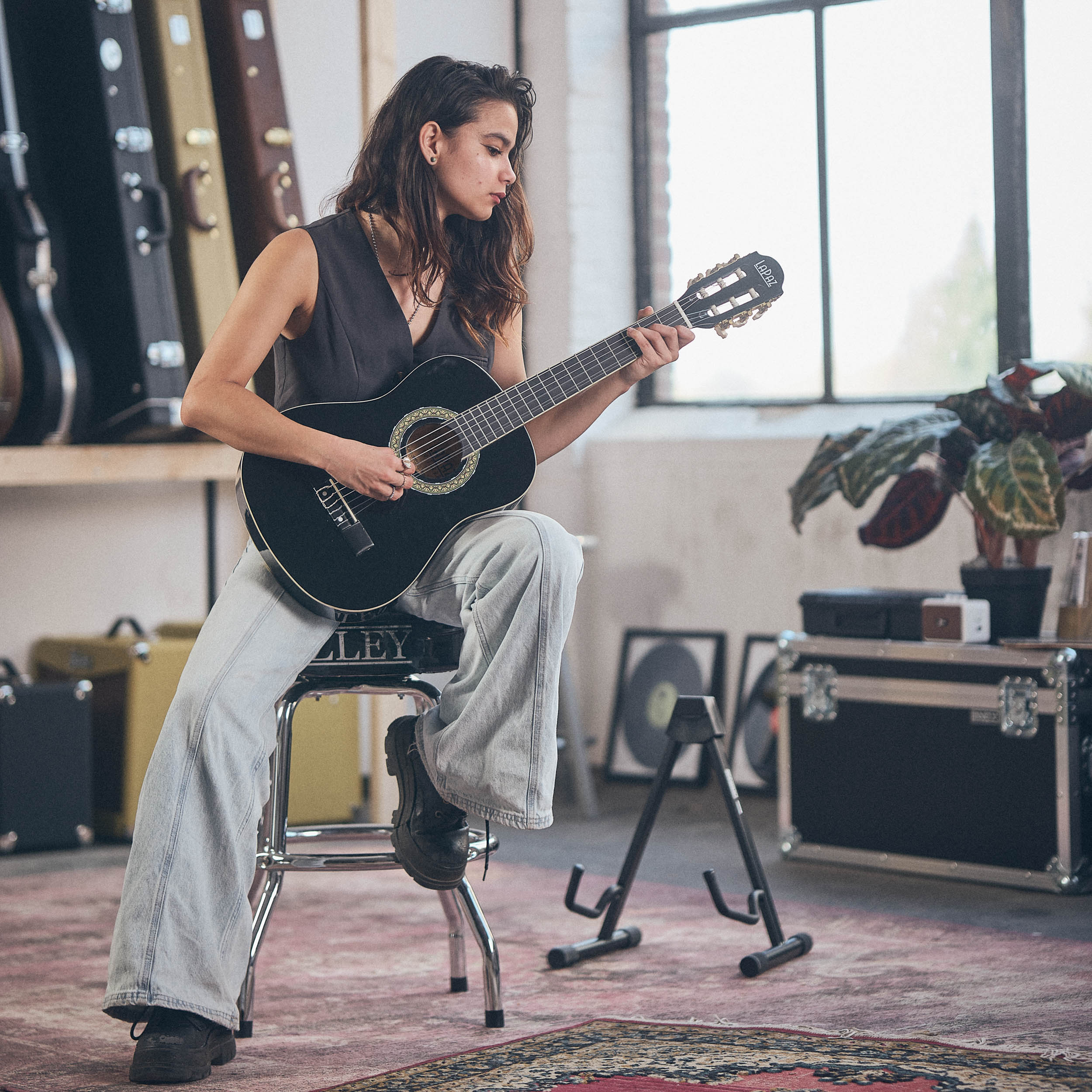 Woman playing black guitar in a room with guitar cases and plants.