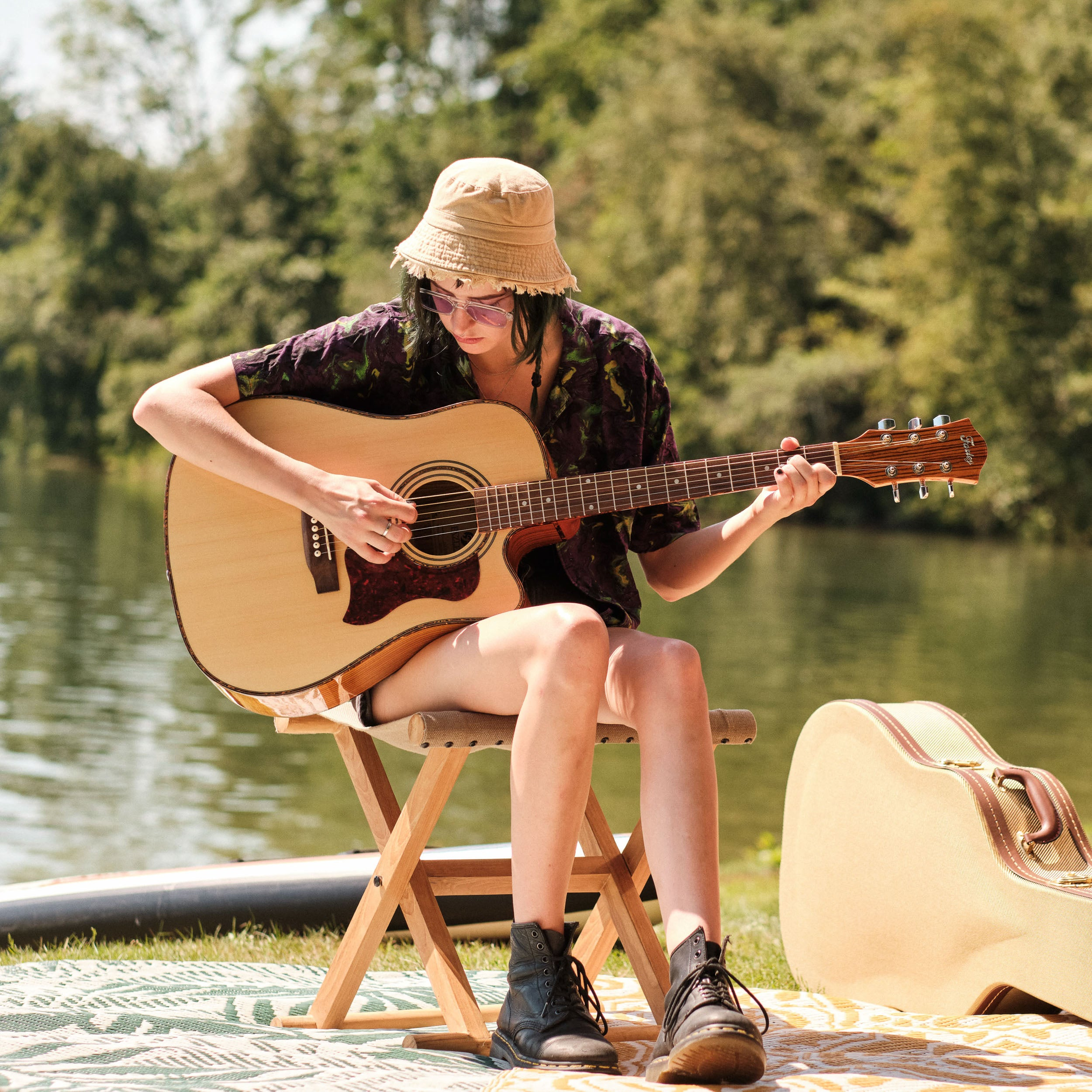 A person sits on a chair playing guitar by the water with a guitar case.