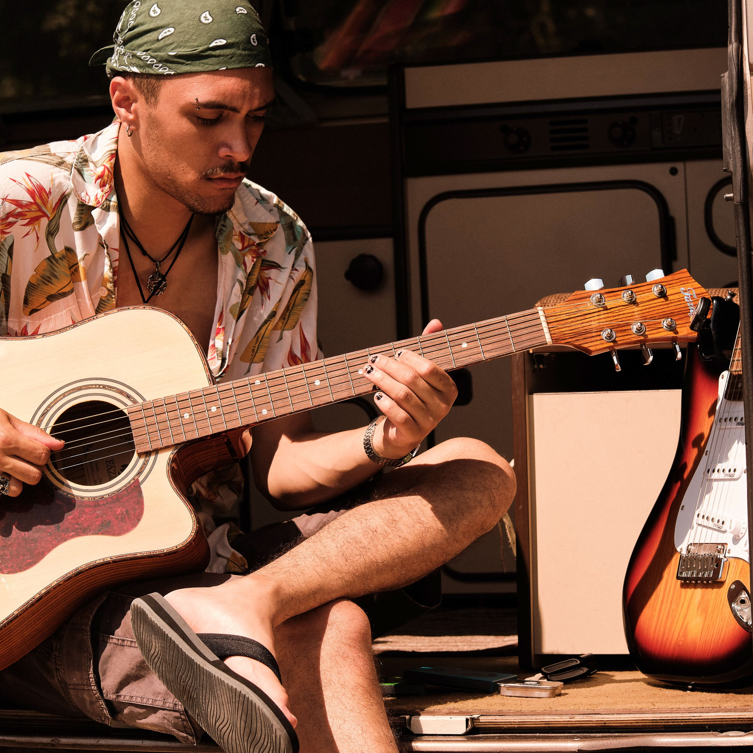 Man in bandana plays acoustic guitar in a van with electric guitar beside him.