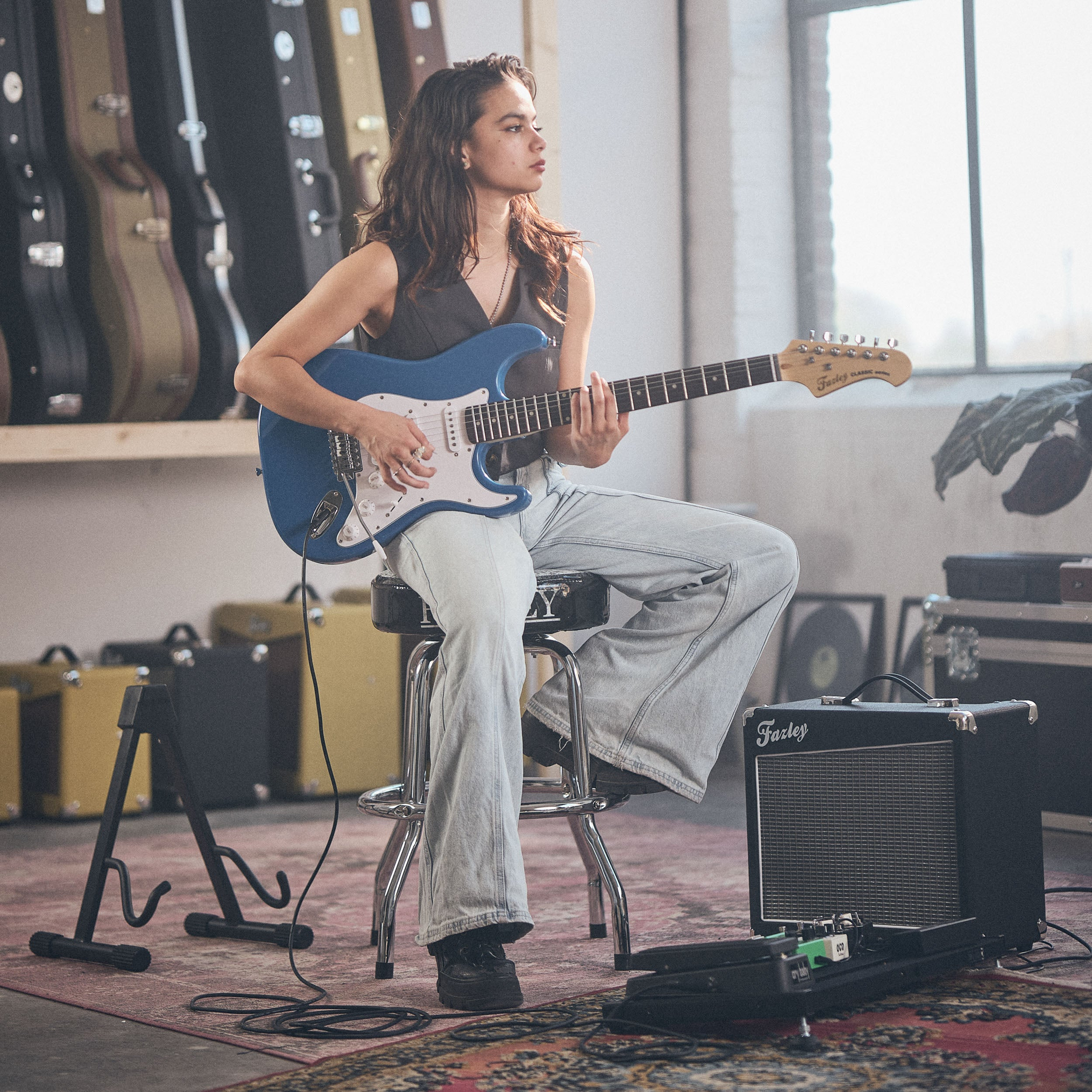 Een vrouw zit op een kruk en speelt op een blauwe elektrische gitaar. Gitaarkoffers op de achtergrond.