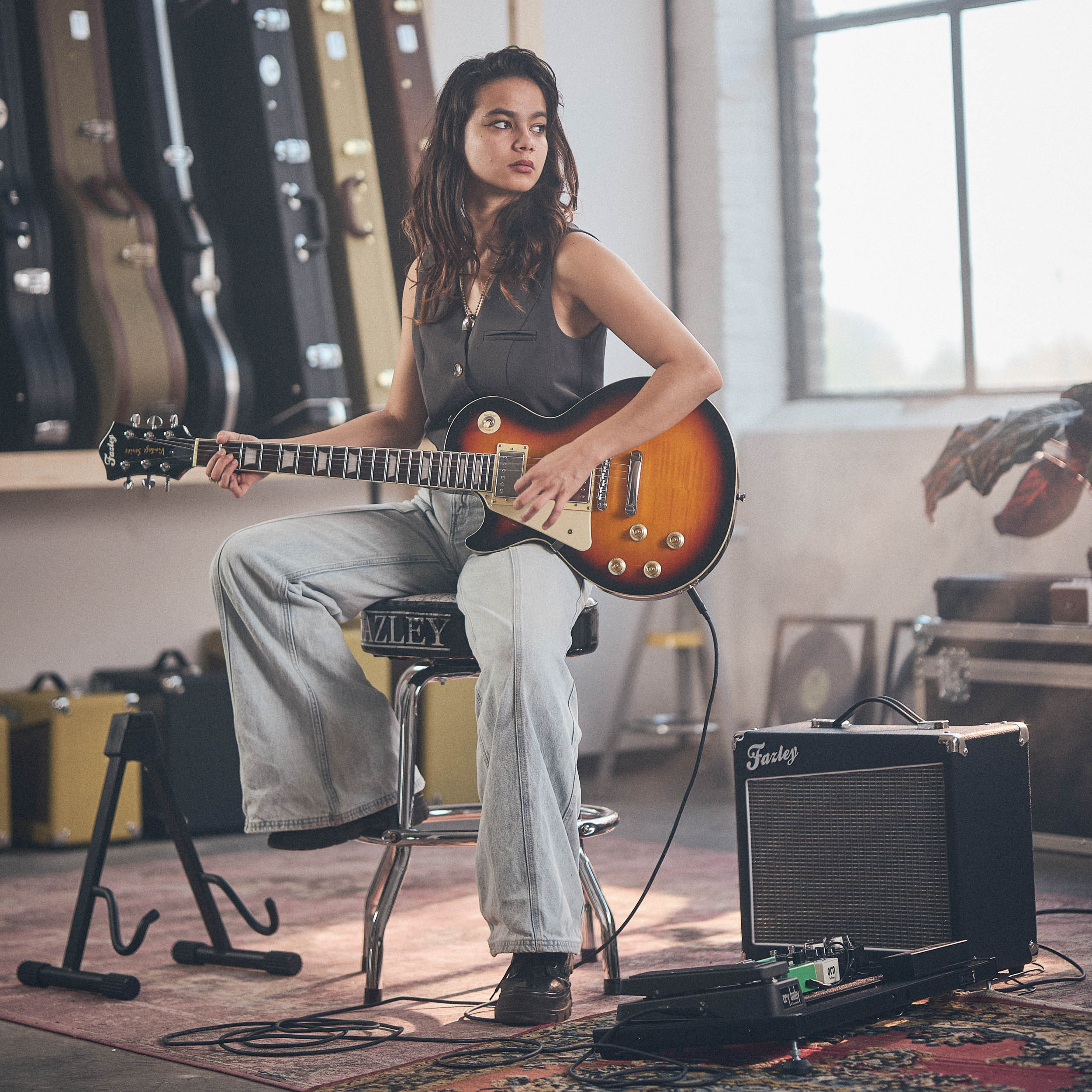 Een vrouw zit op een kruk en speelt gitaar, met gitaren op de achtergrond.