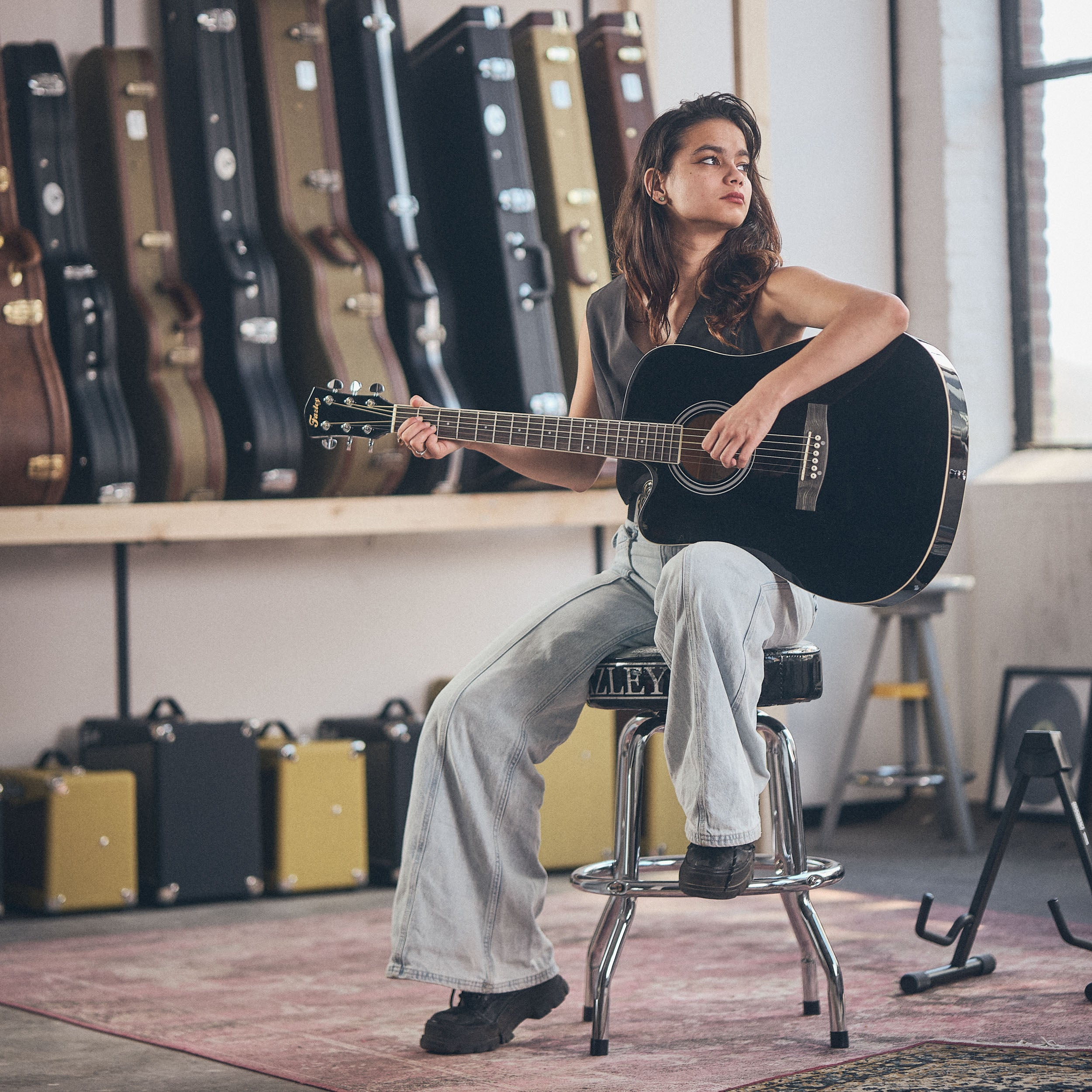 Een vrouw zit op een kruk en speelt gitaar. Veel gitaarkoffers op de achtergrond.