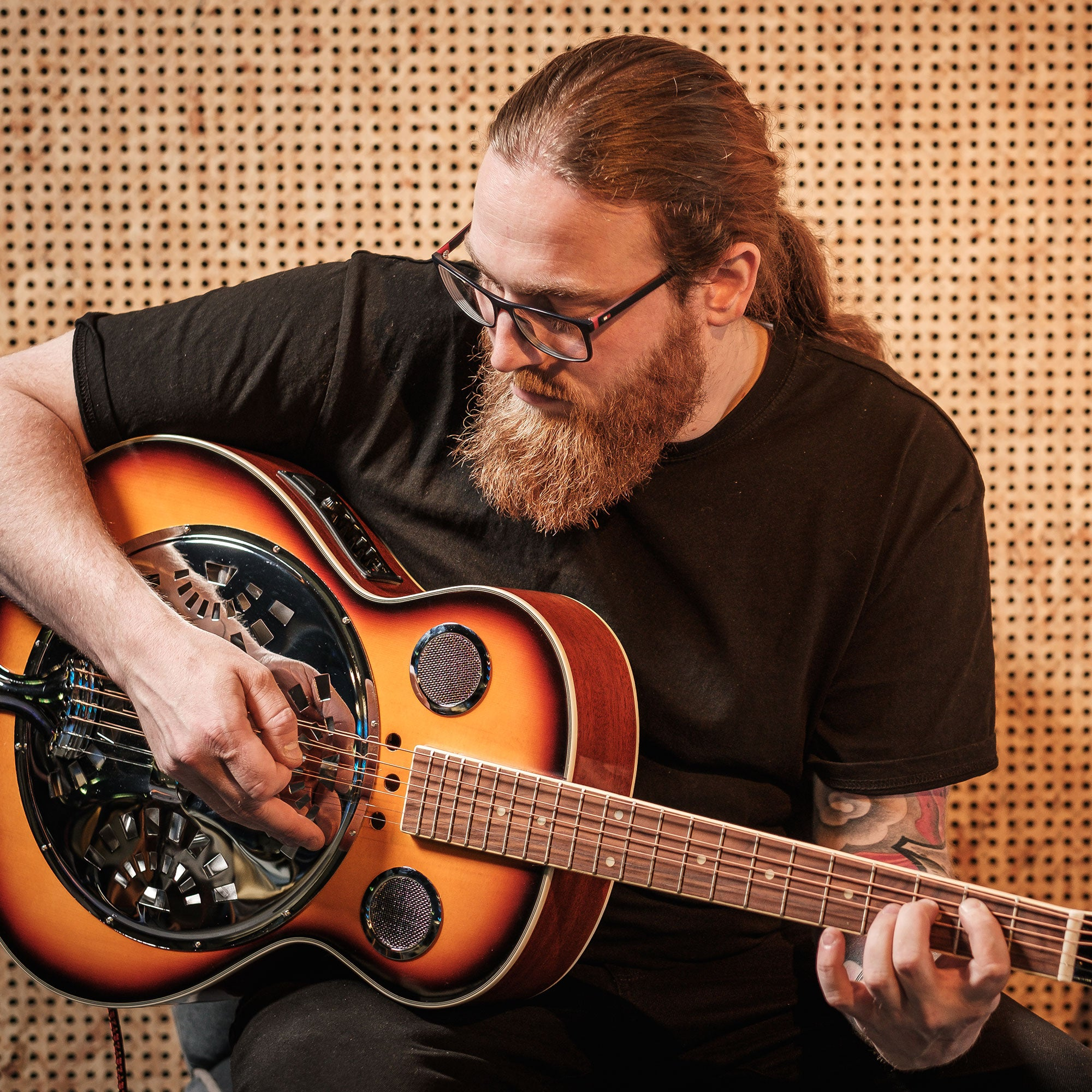 Man met bril en baard speelt op een bruine gitaar voor een houten wand.