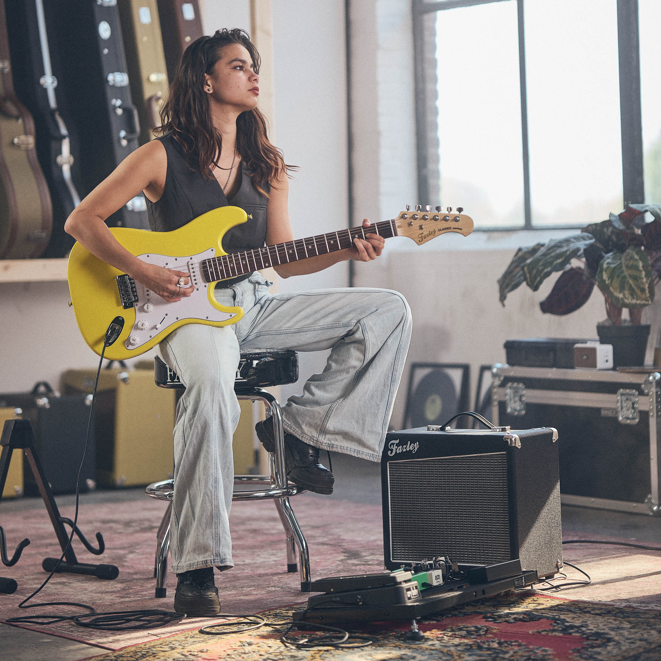 Een vrouw speelt op een gele elektrische gitaar, zittend op een kruk, studio setting.