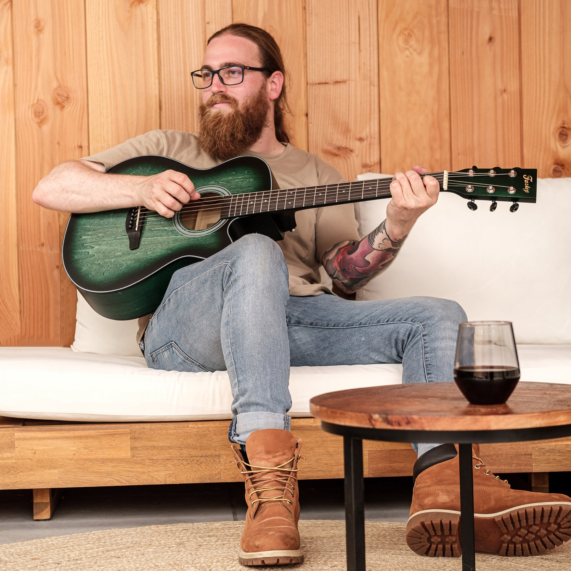 Man met baard speelt groene gitaar op bank, glas wijn ernaast.