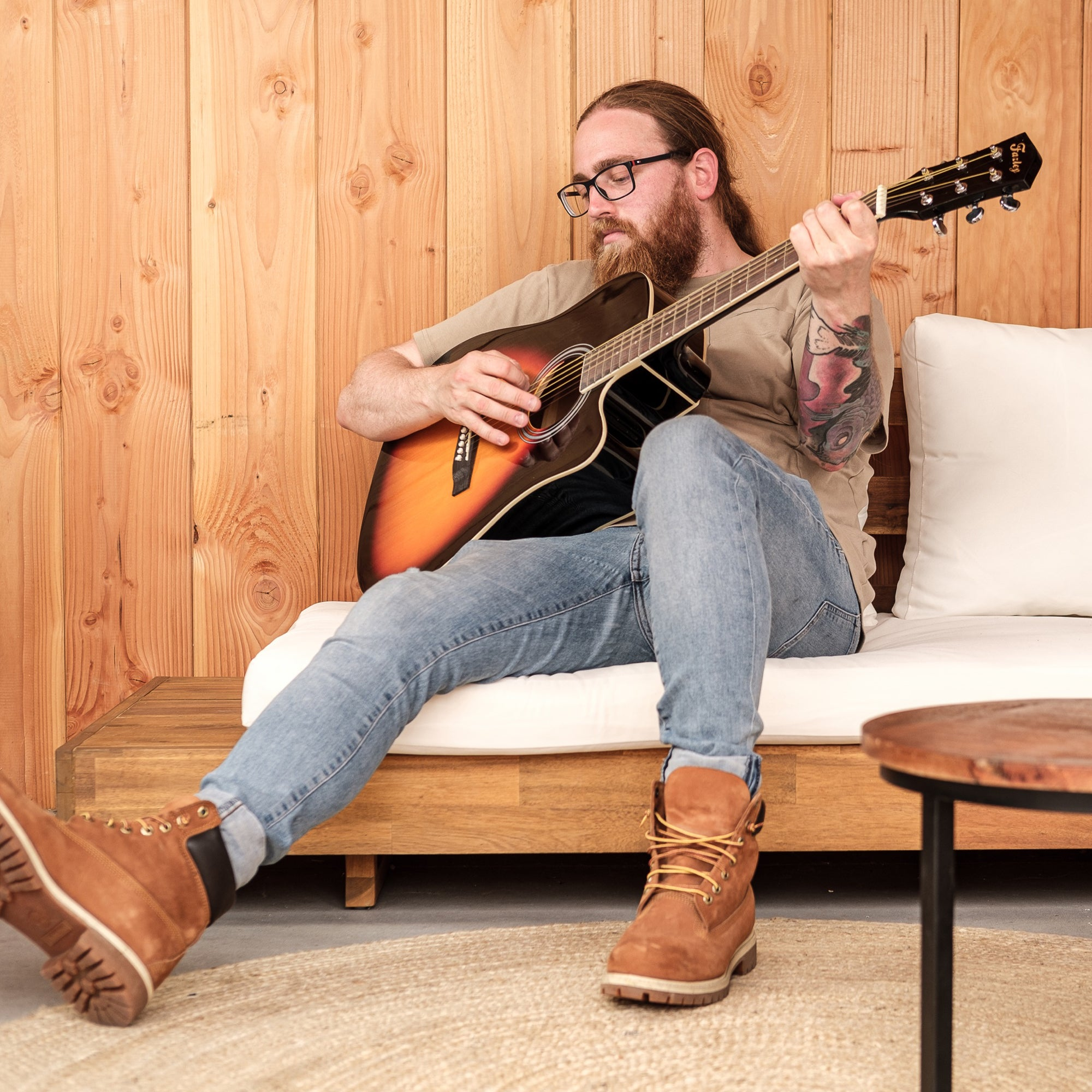 Man met baard speelt akoestische gitaar op bank voor houten lambrisering.