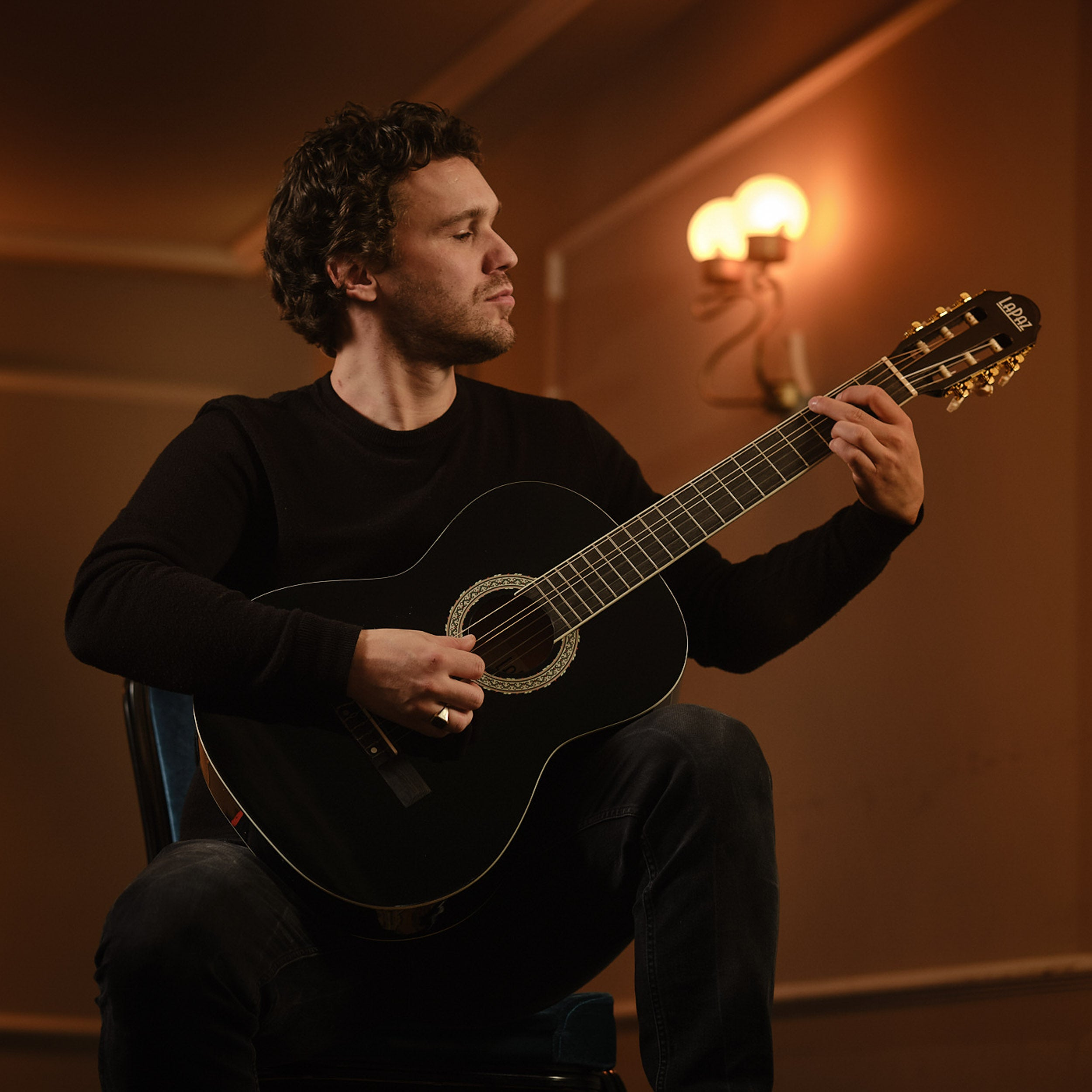 Een man met donker haar speelt op een zwarte gitaar, verlicht door warm licht.