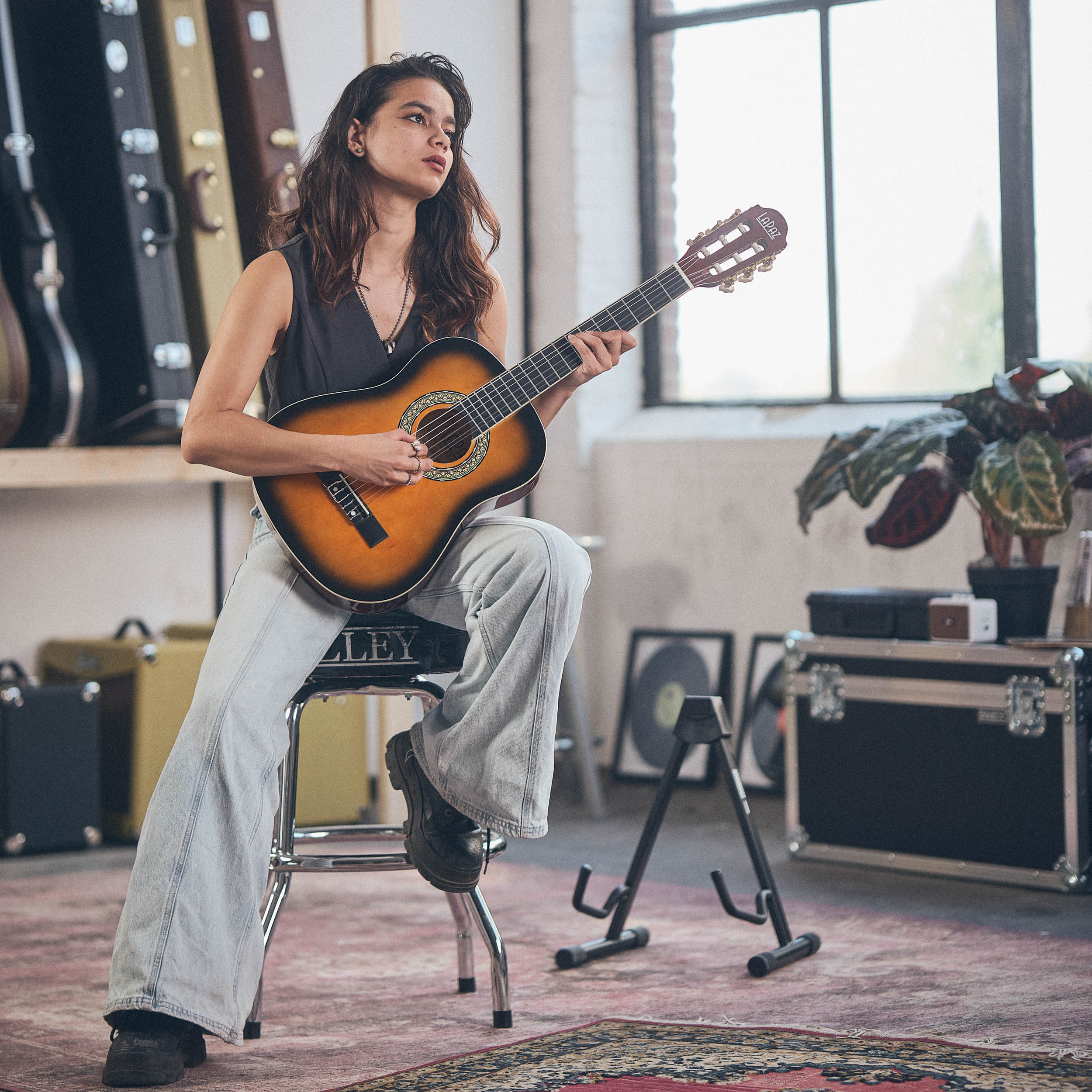 Een vrouw zit op een kruk en speelt gitaar. Diverse gitaarkoffers staan op de achtergrond.