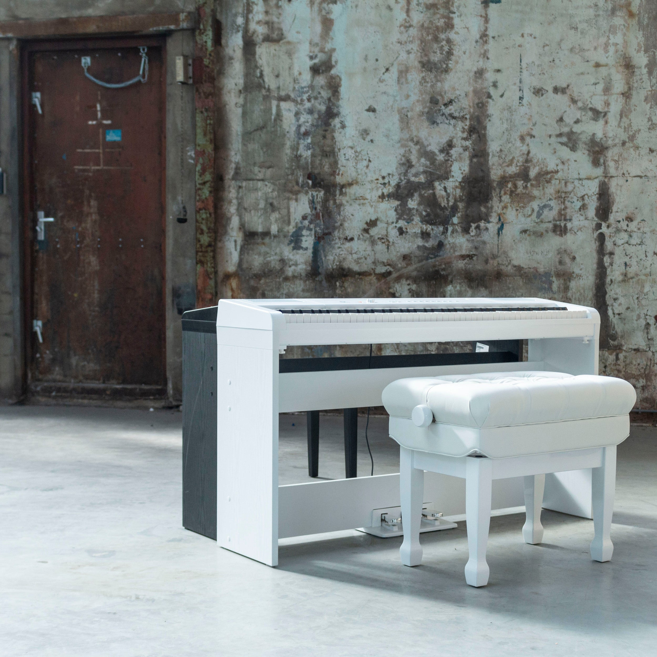 Een witte digitale piano met kruk staat voor een roestige deur en betonnen muur.