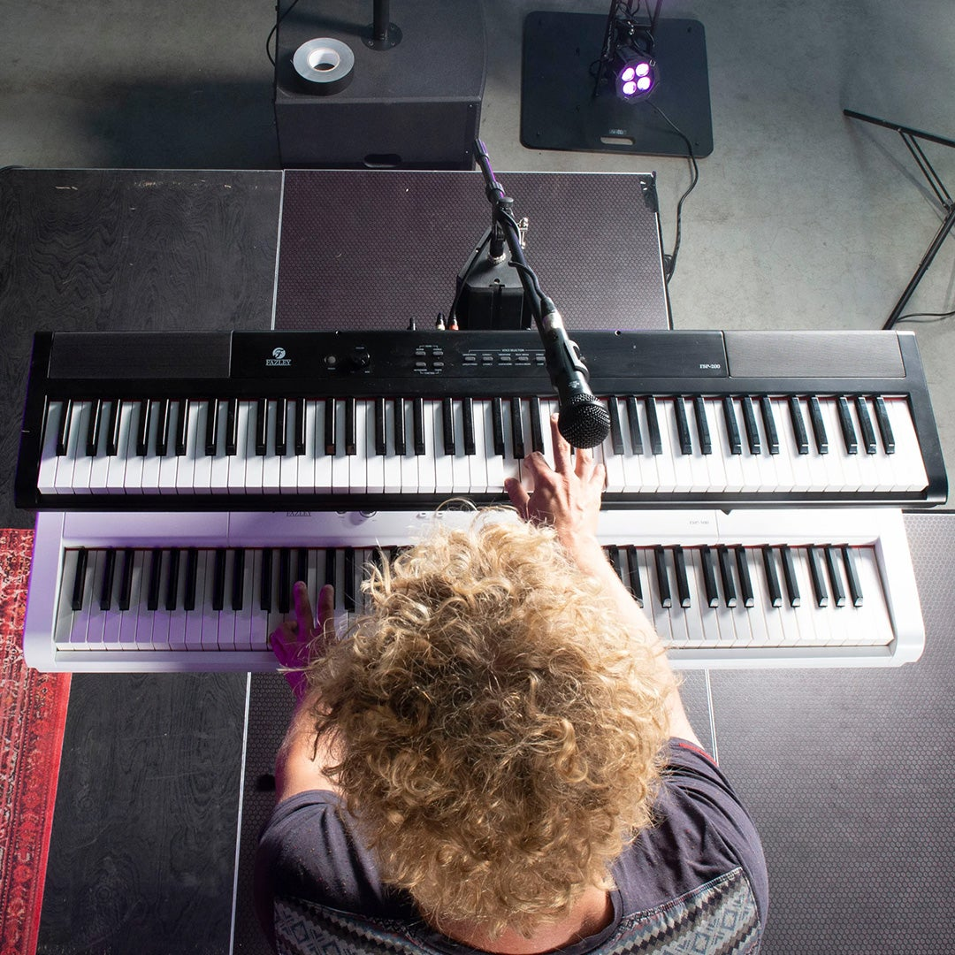Persoon met krullend haar speelt op twee piano's, één zwart, één wit.