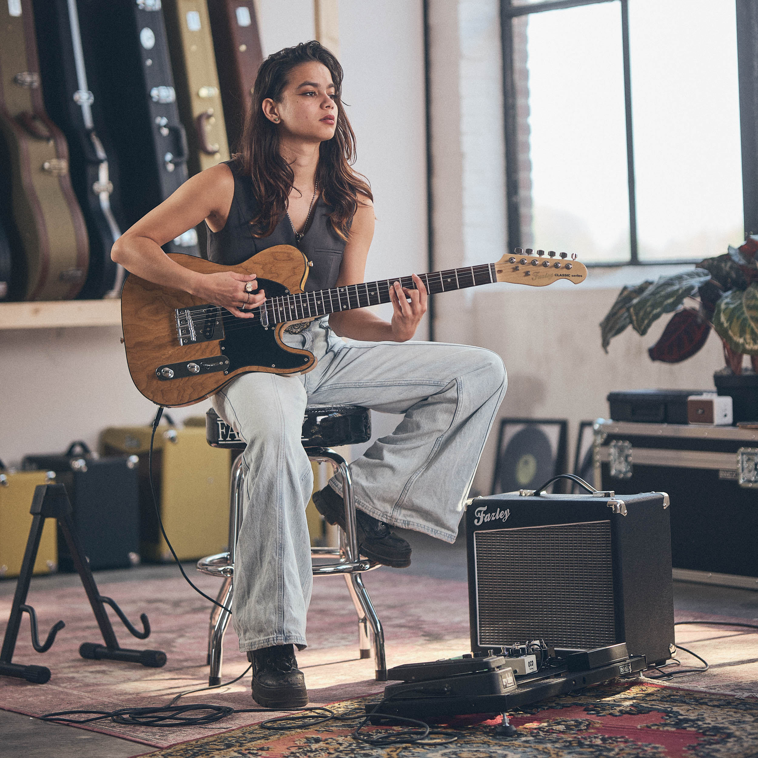 Jonge vrouw zit op krukje gitaar spelend in kamer met gitaarkoffers.