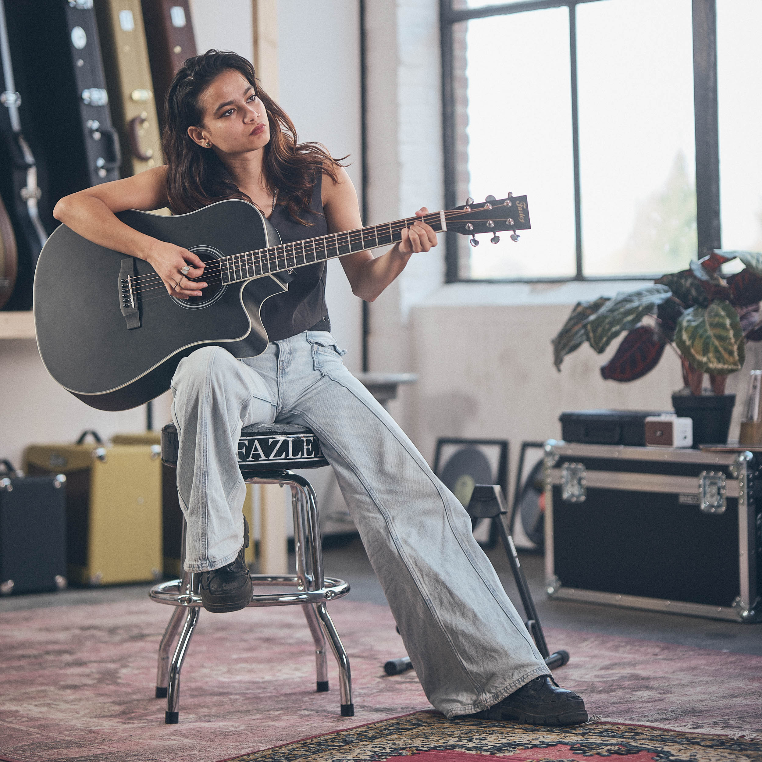 Een vrouw met donker haar speelt op een zwarte akoestische gitaar, zittend op een kruk.