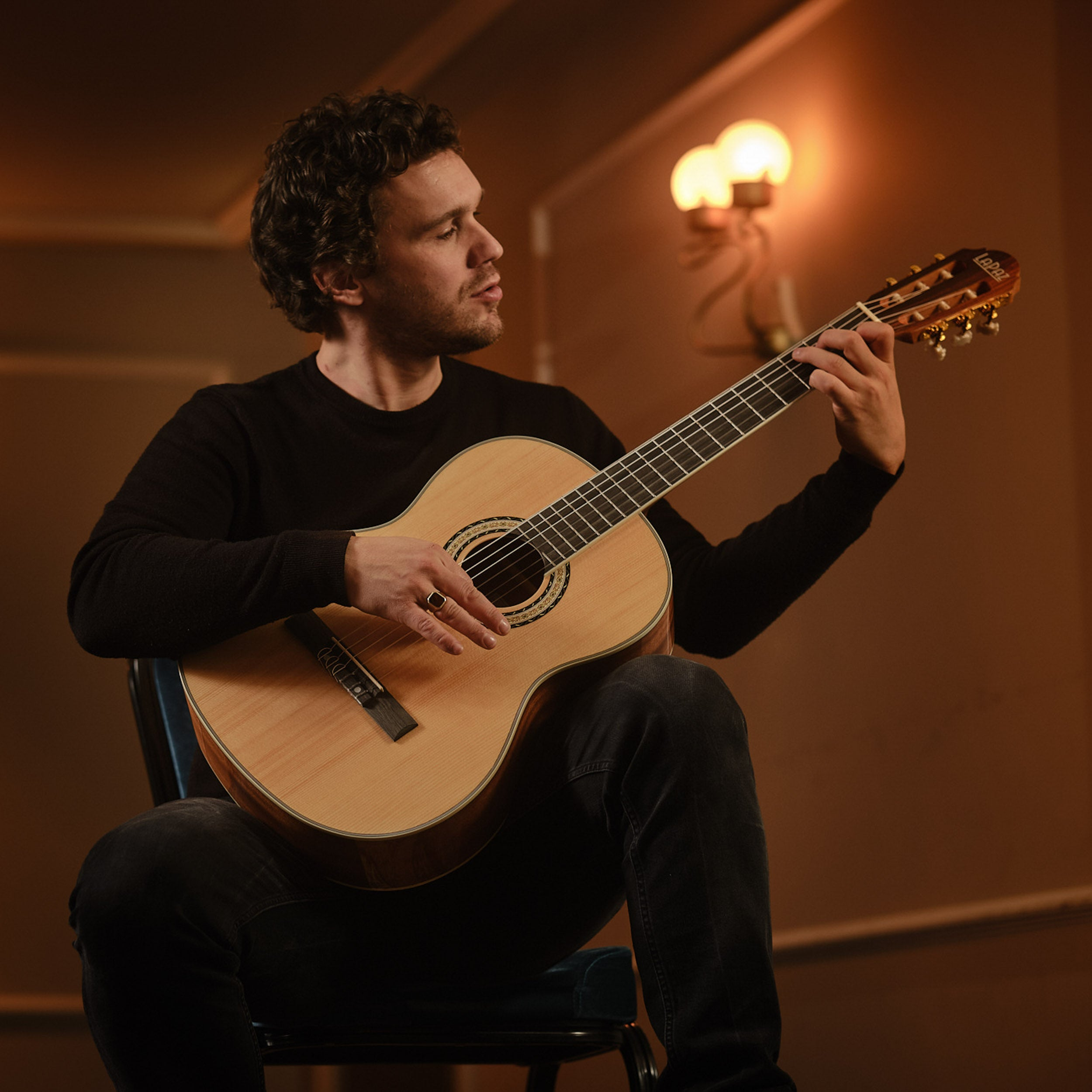 Een man met donker haar speelt akoestische gitaar, zittend op een stoel.
