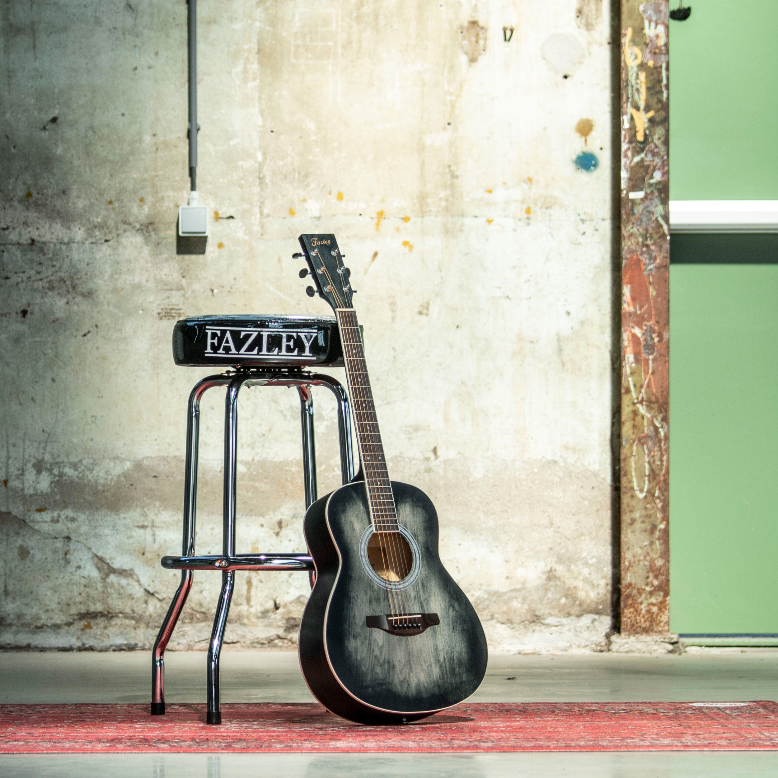 Een donkere akoestische gitaar leunt tegen een kruk met "FAZLEY" erop.