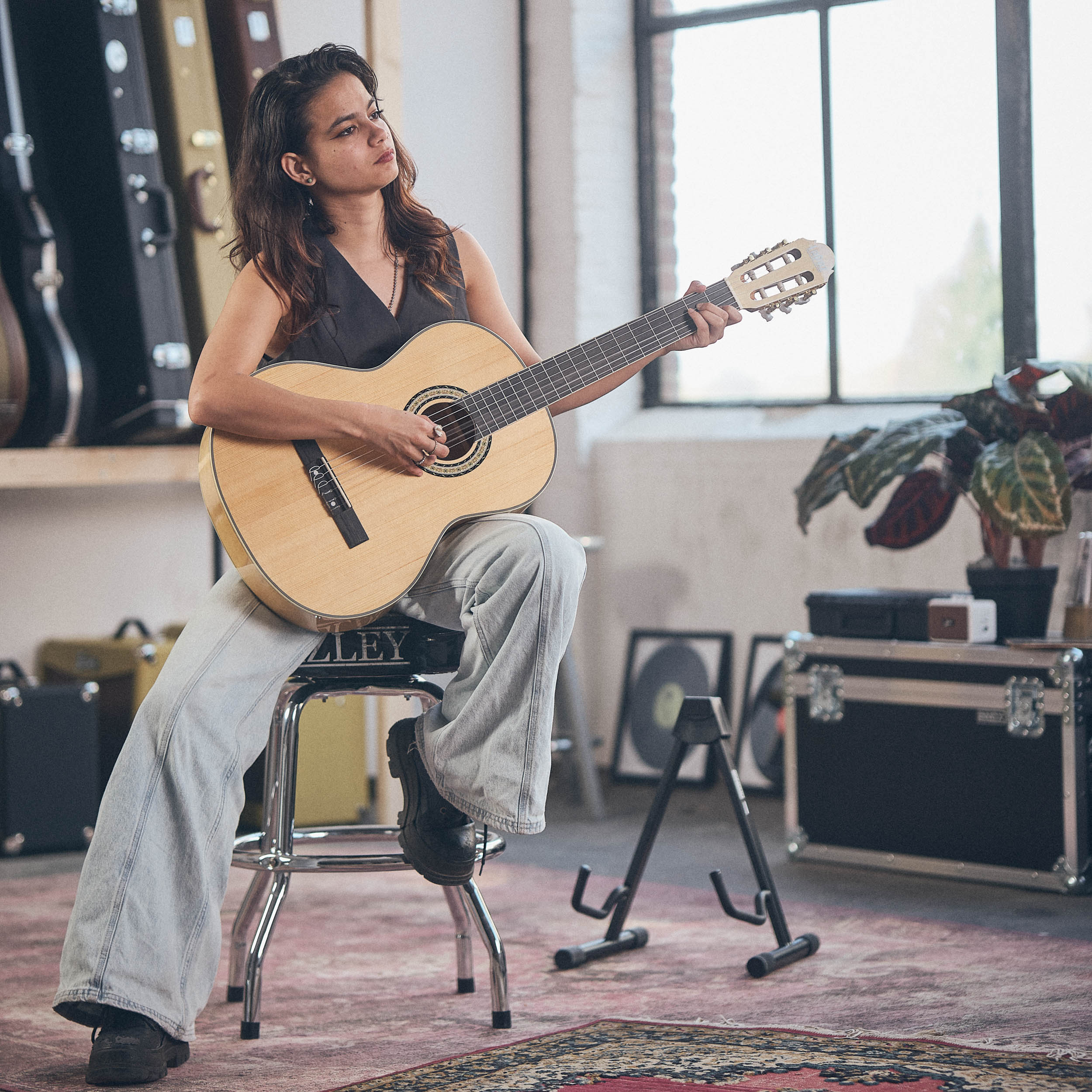Een vrouw zit op een krukje gitaar te spelen in een kamer met gitaarkoffers.