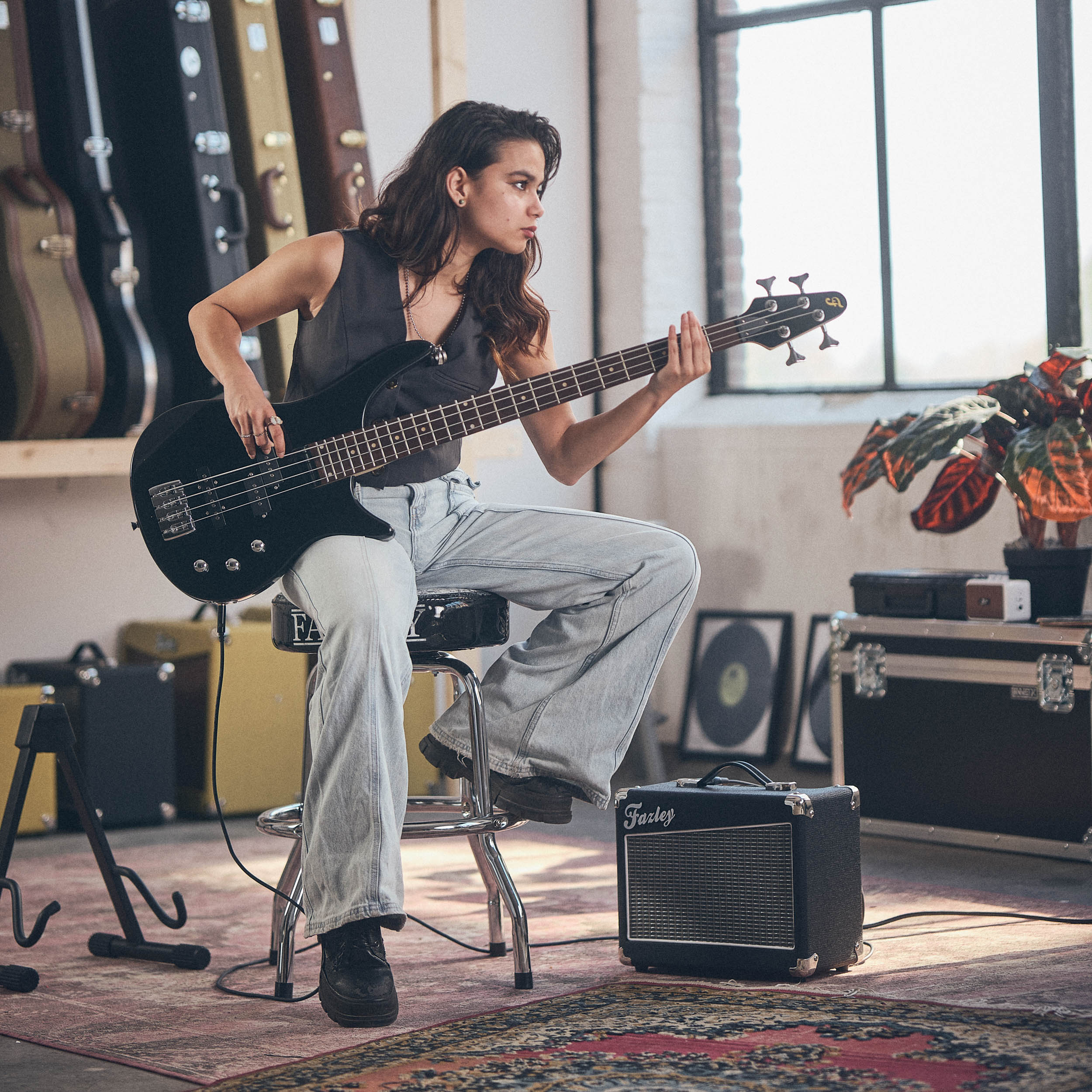 Een vrouw zit op een kruk en bespeelt een zwarte basgitaar in een kamer vol gitaarkoffers.