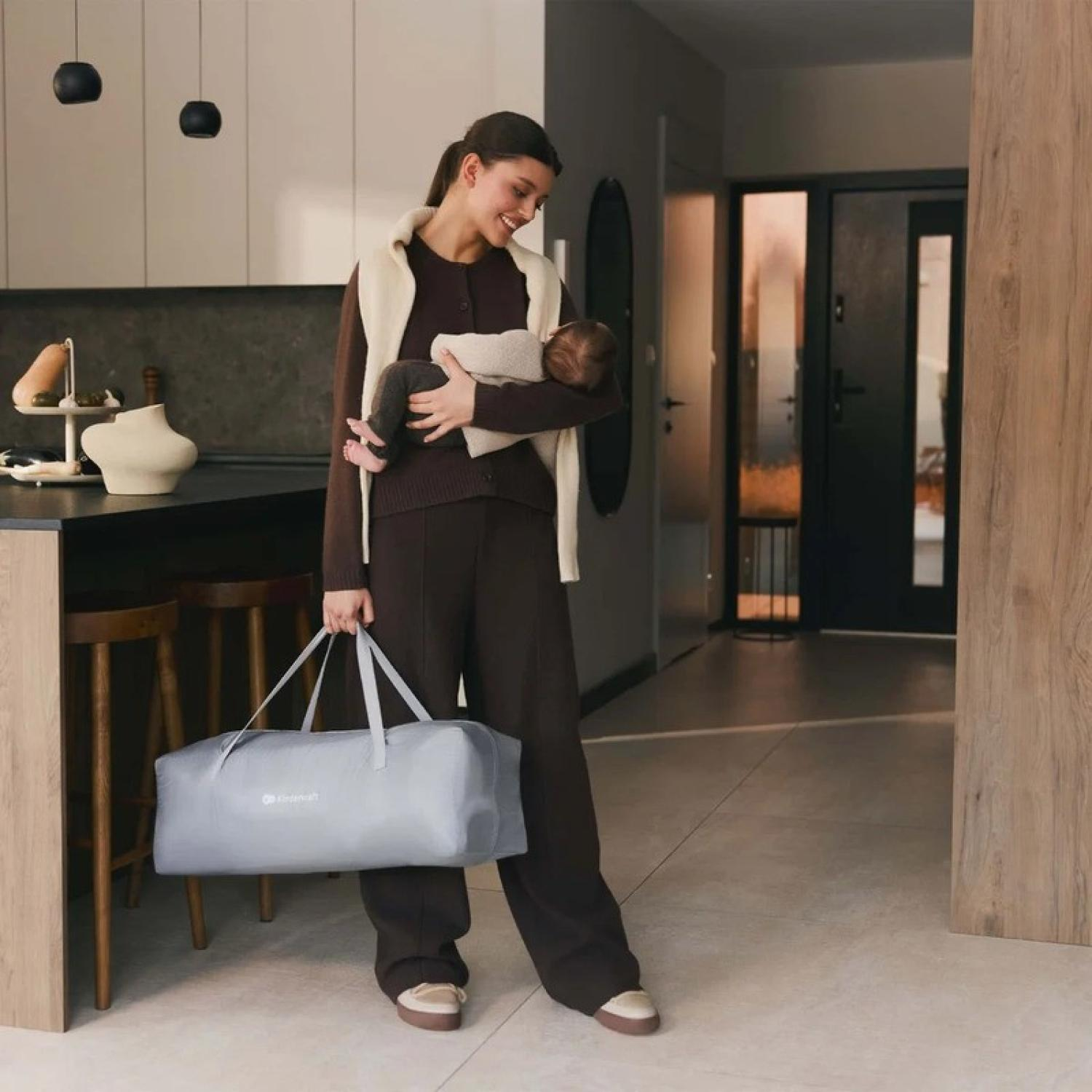 Eine Frau trägt ein Baby und eine graue Tasche in einer modernen Küche. Holzakzente im Hintergrund.